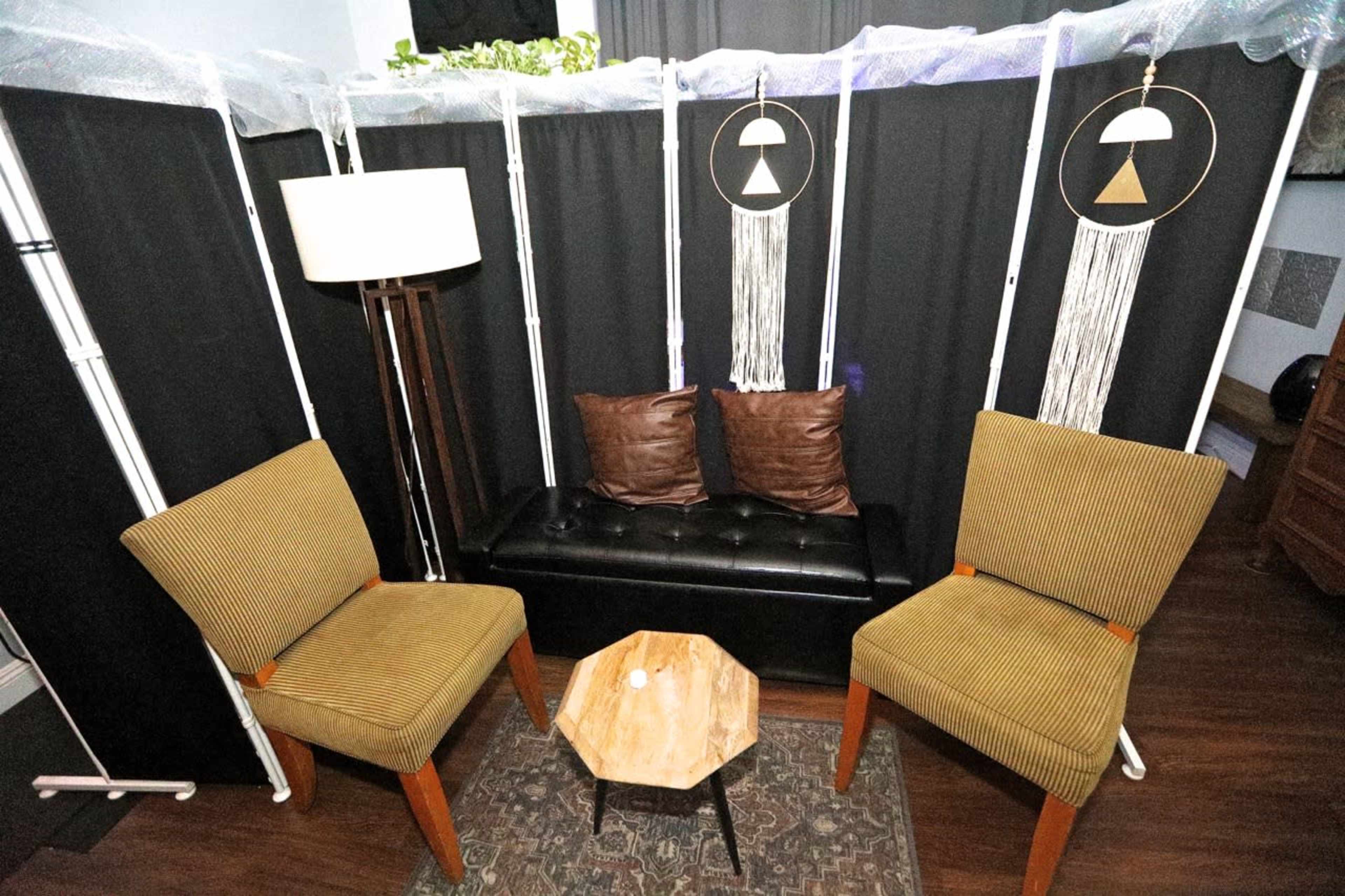 A small, enclosed seating area with two striped chairs, a black loveseat, a round wooden coffee table, and decorative hanging elements.