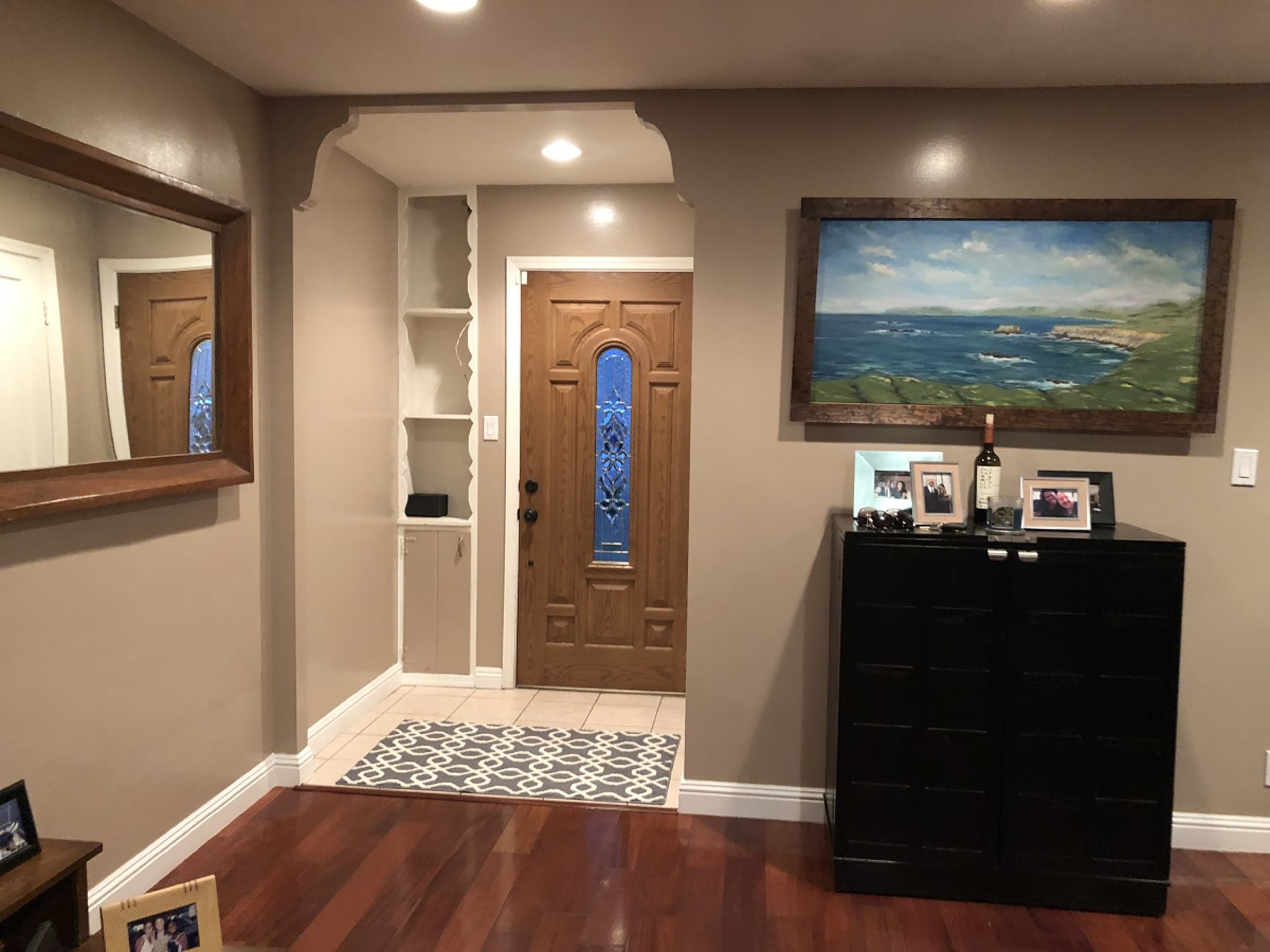 A living room entrance with a wooden door, a decorative painting on the wall, a mirror, and a dark cabinet, all situated on a hardwood floor.