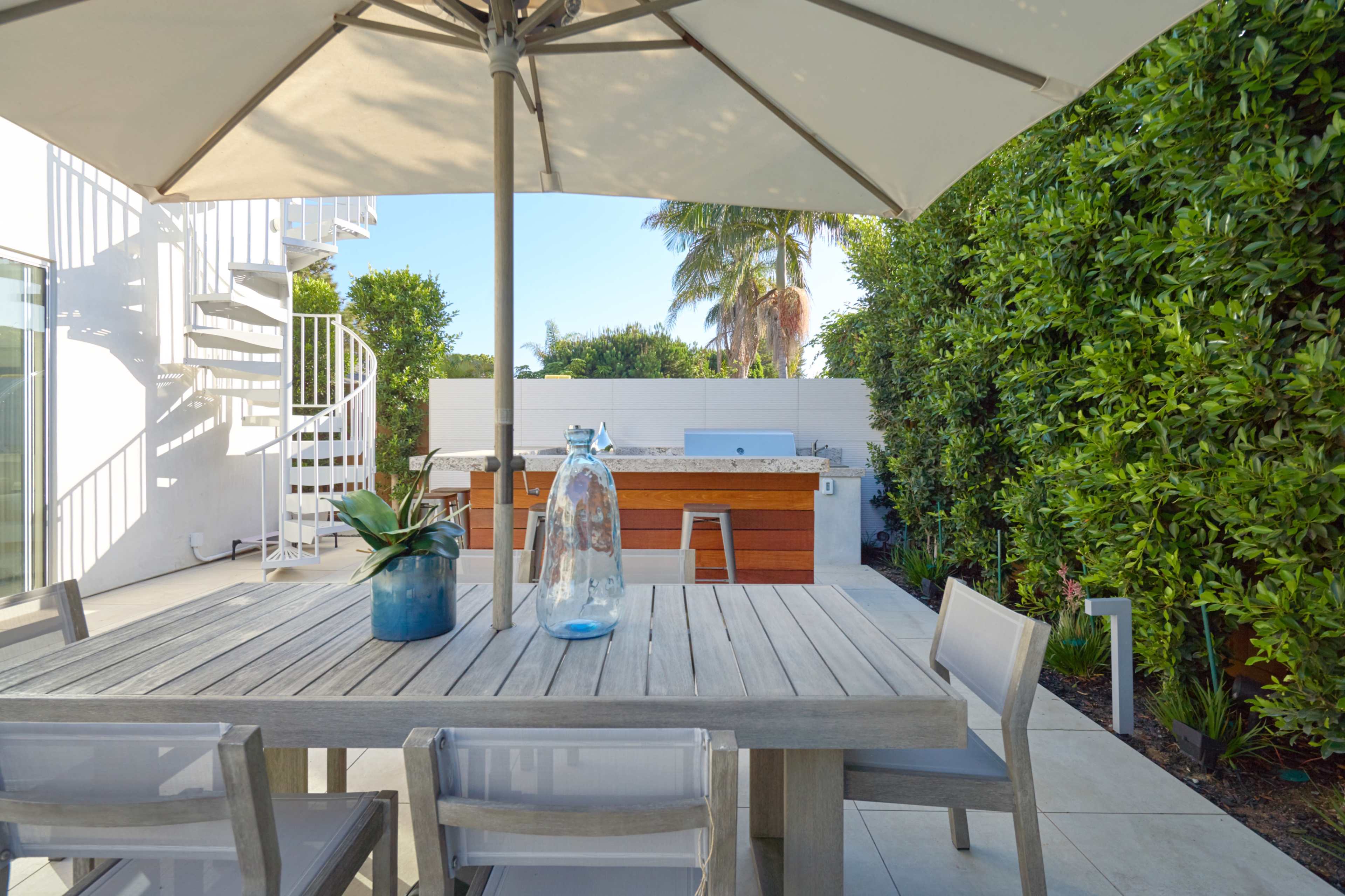A wooden table with clear chairs is set under a large umbrella, with a potted plant and water jug on the table, overlooking an outdoor kitchen surrounded by greenery.