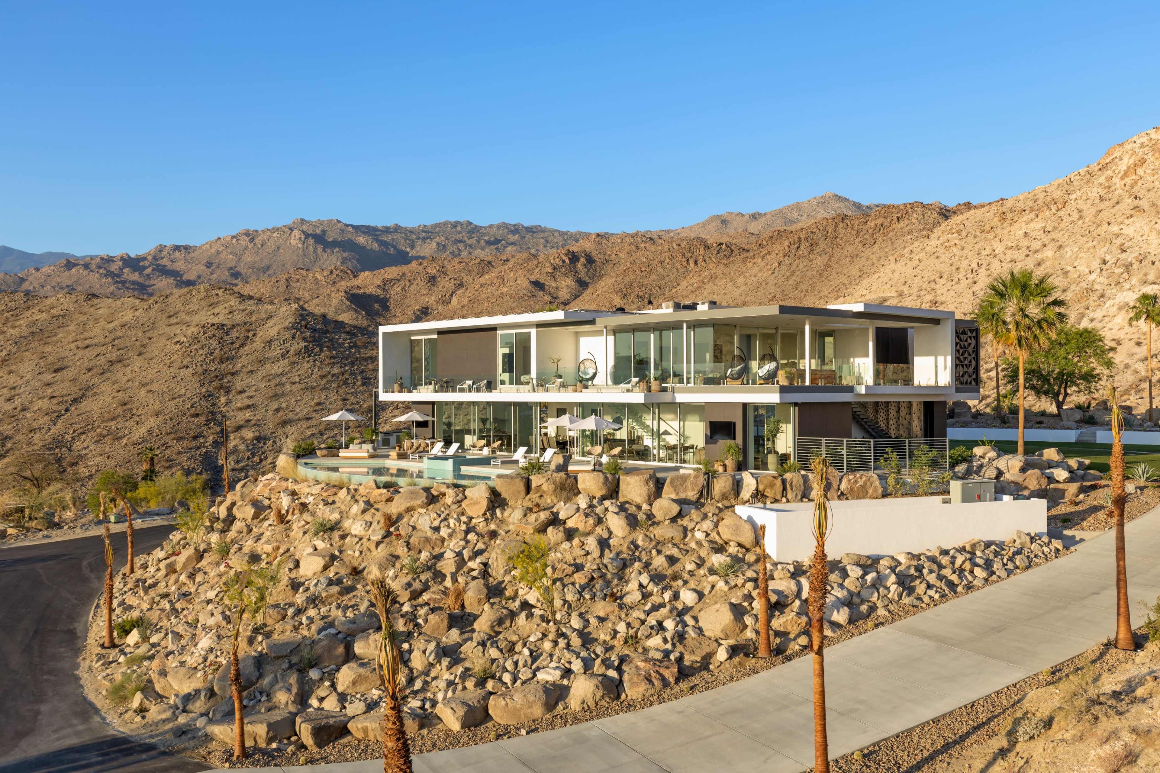A modern two-story house is perched on a rocky slope, surrounded by desert landscapes and distant mountains.