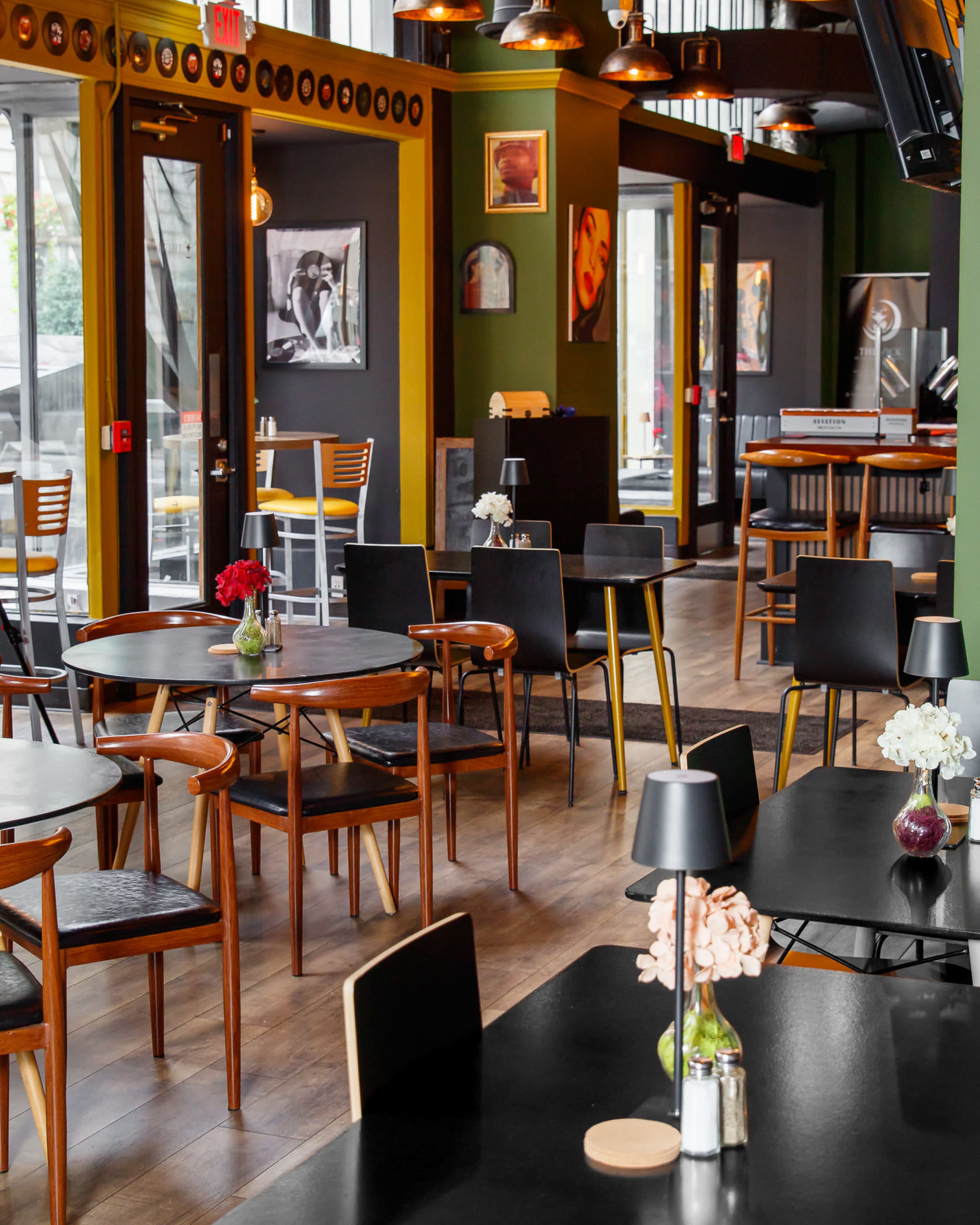 The image shows a modern café interior with wooden and black tables, chairs, and decorative flowers on each table, along with large windows and artistic wall decor.