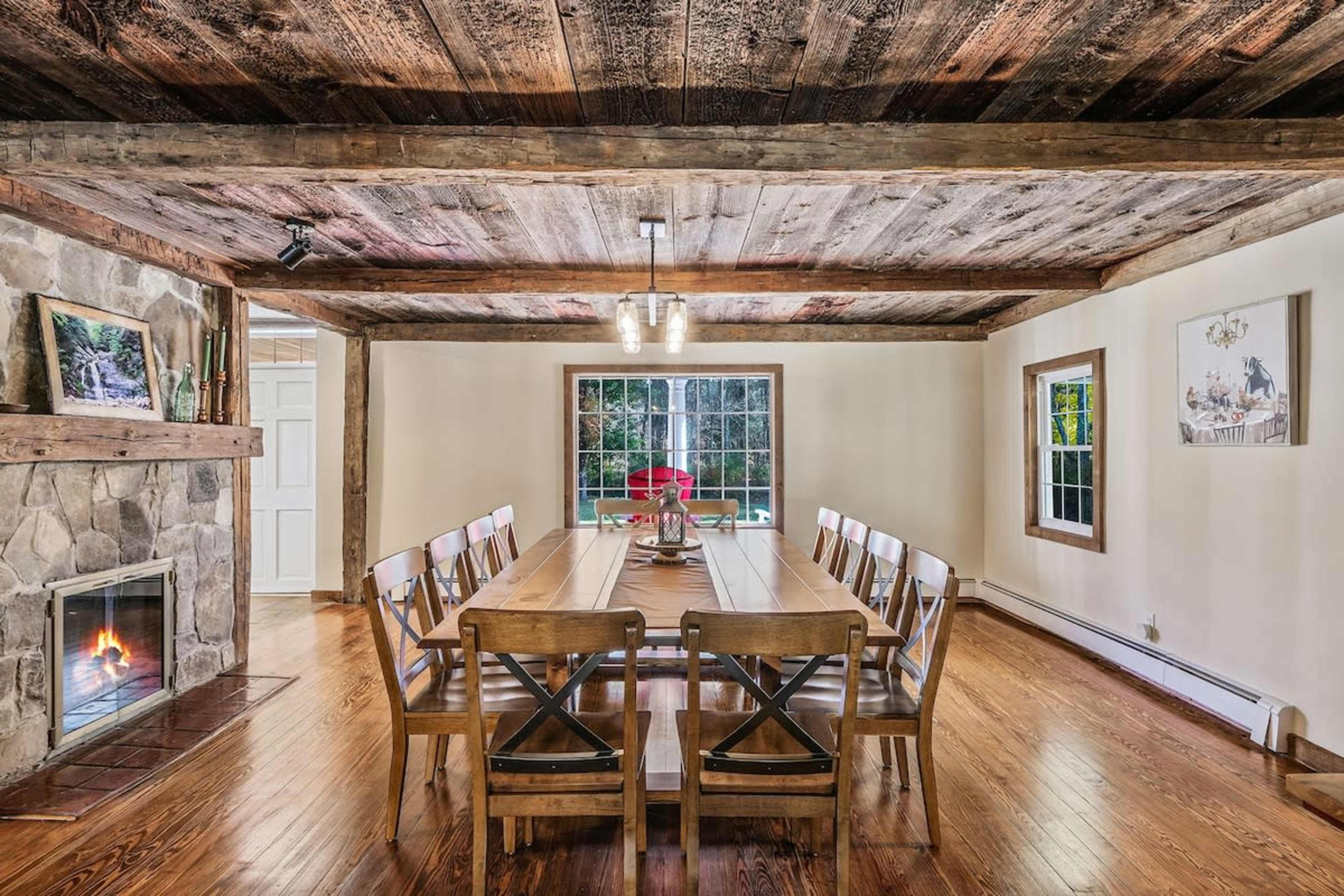 A large wooden dining table with twelve chairs is positioned in a rustic room featuring exposed beams and a stone fireplace.