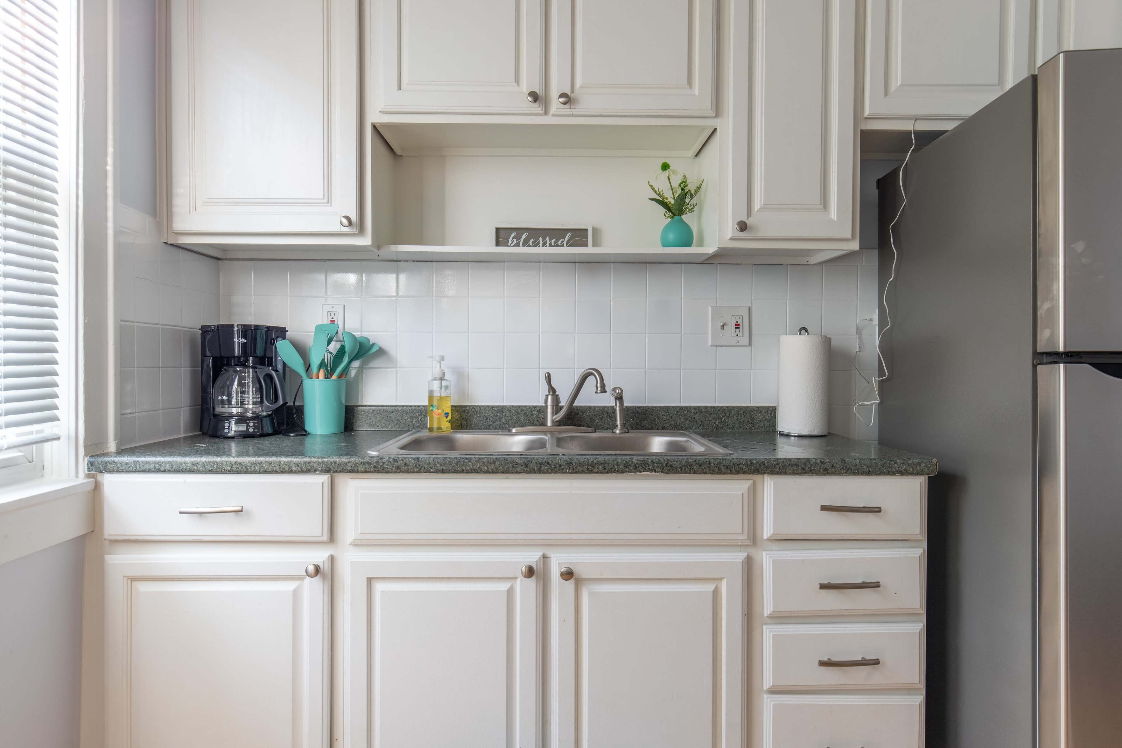 A clean kitchen counter features a coffee maker, a stainless steel refrigerator, a sink with dish soap, and neatly organized utensils in a turquoise holder.