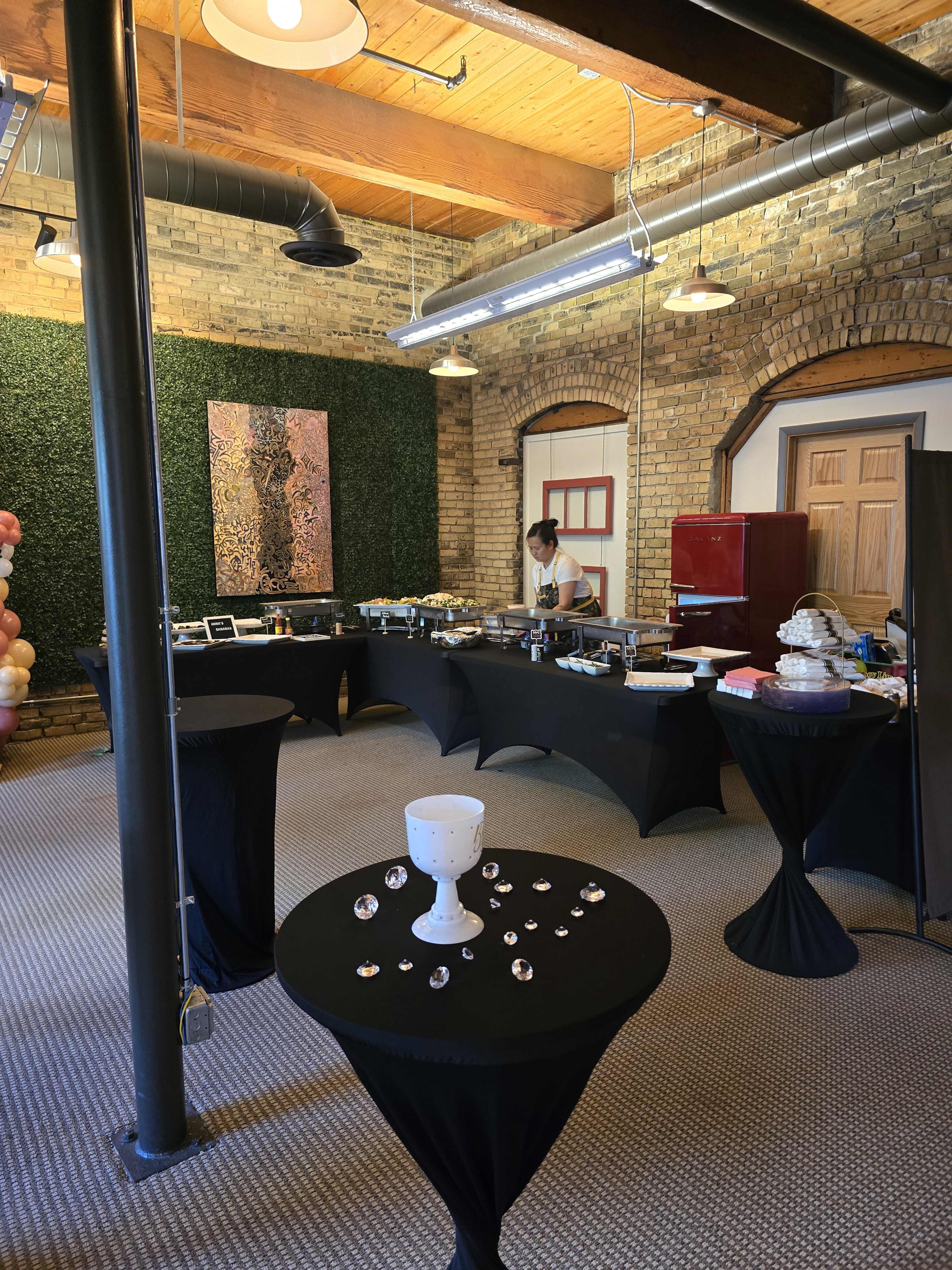 A spacious event room with black tablecloths, a buffet setup, and a person preparing food against a backdrop of brick walls and decorative greenery.