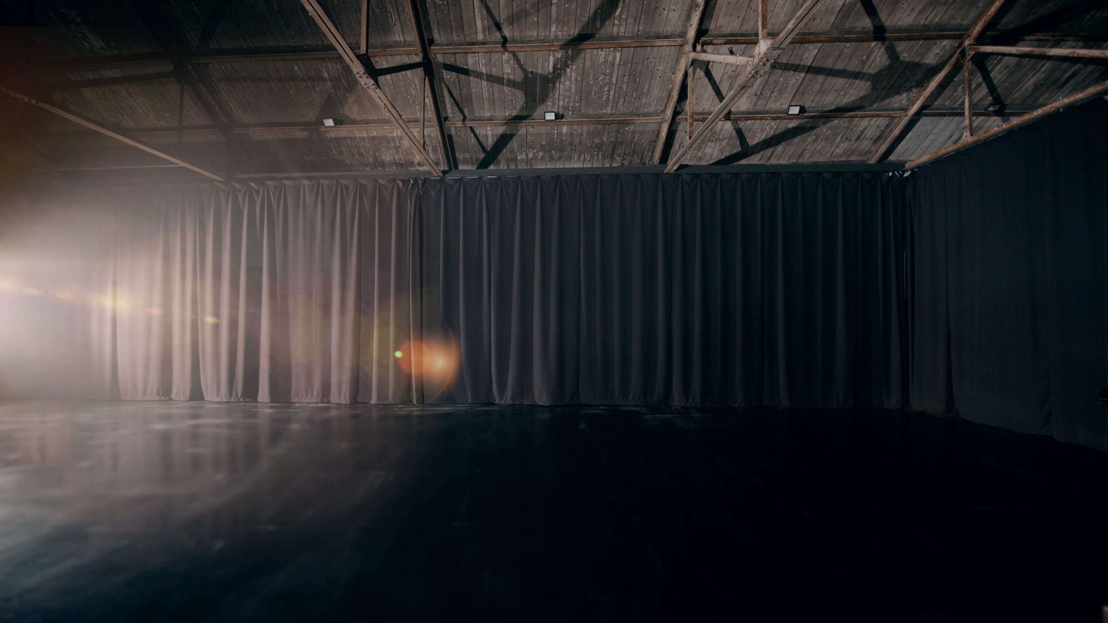 The image shows a dimly lit indoor space with a dark curtain and a spotlight casting a soft glow on the floor.