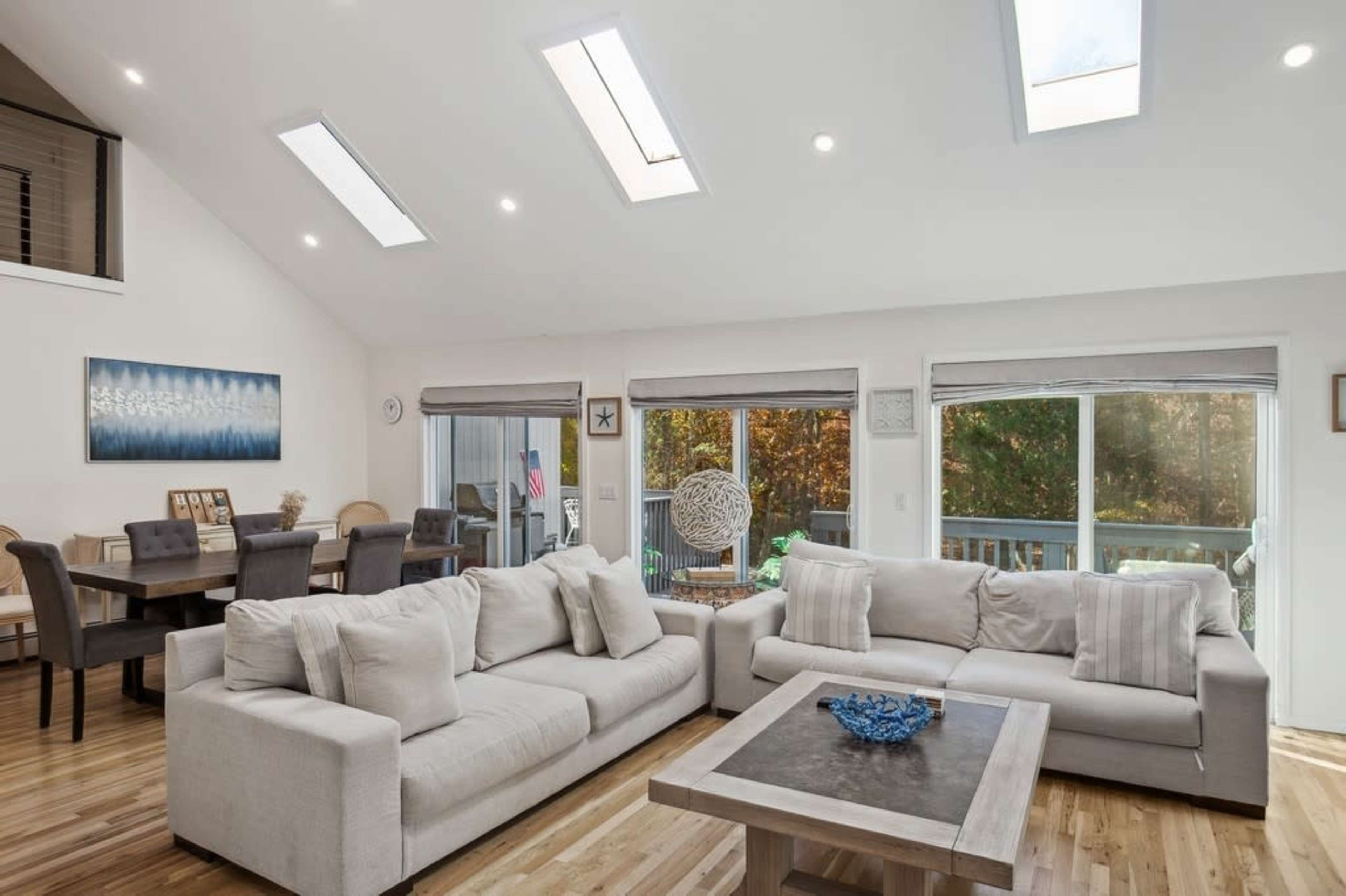 The image shows a modern living room with a light-colored sectional sofa, a coffee table, and large windows that provide ample natural light.