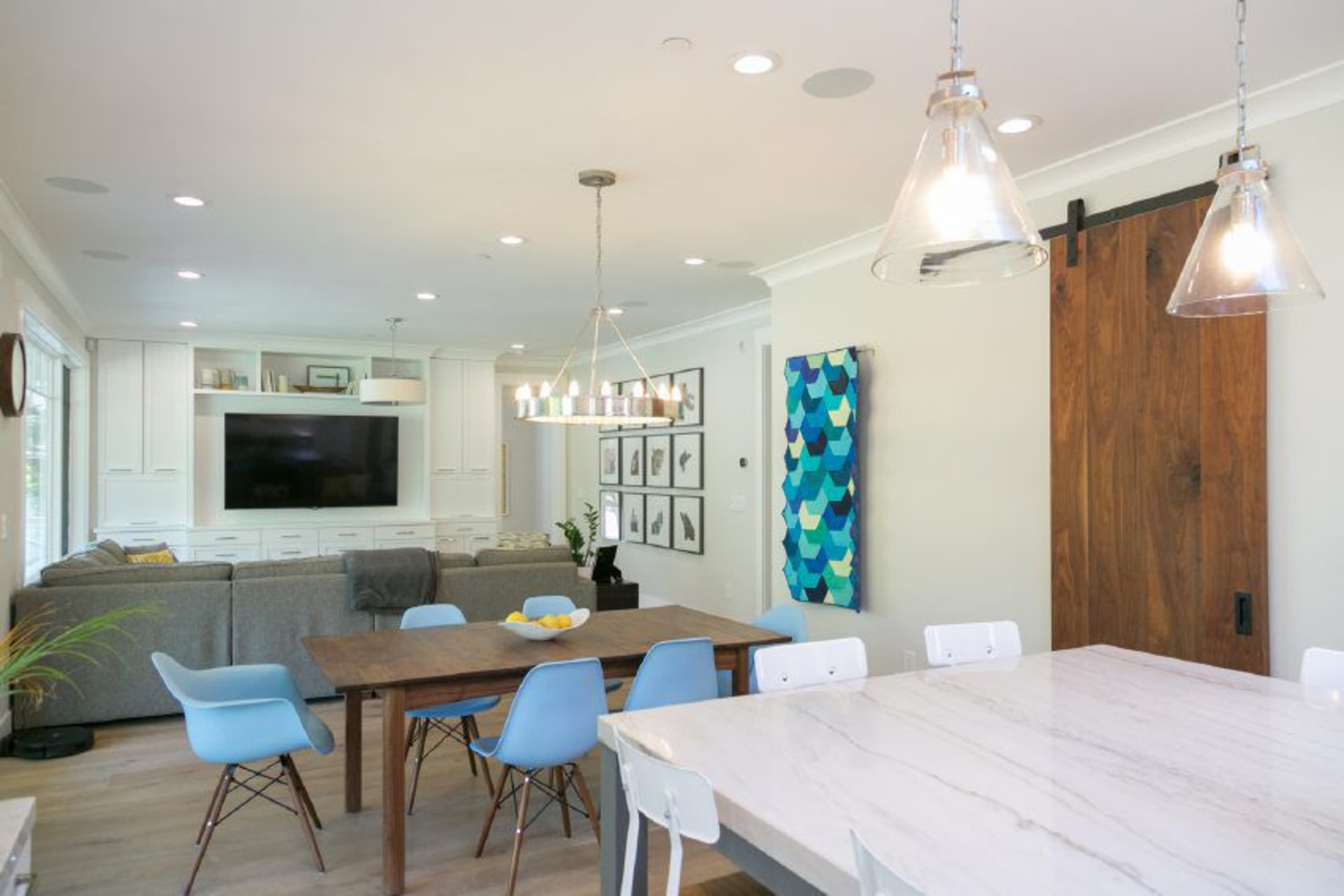 A bright dining area features a wooden table with blue and white chairs, a marble countertop, and a large sliding barn door leading to a living space with a gray sofa and a wall-mounted TV.