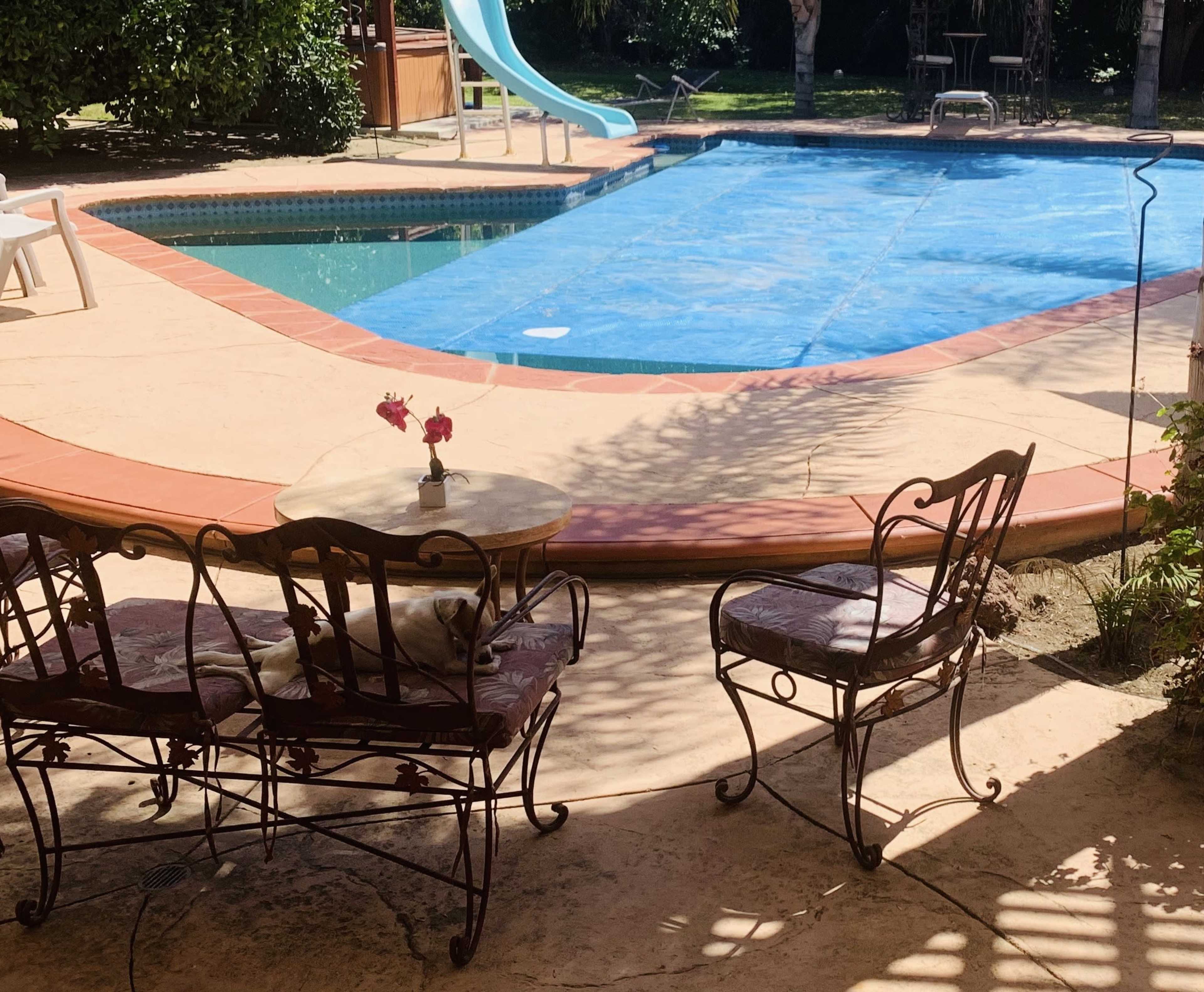 A small table with two chairs sits beside a covered swimming pool in a sunny backyard.