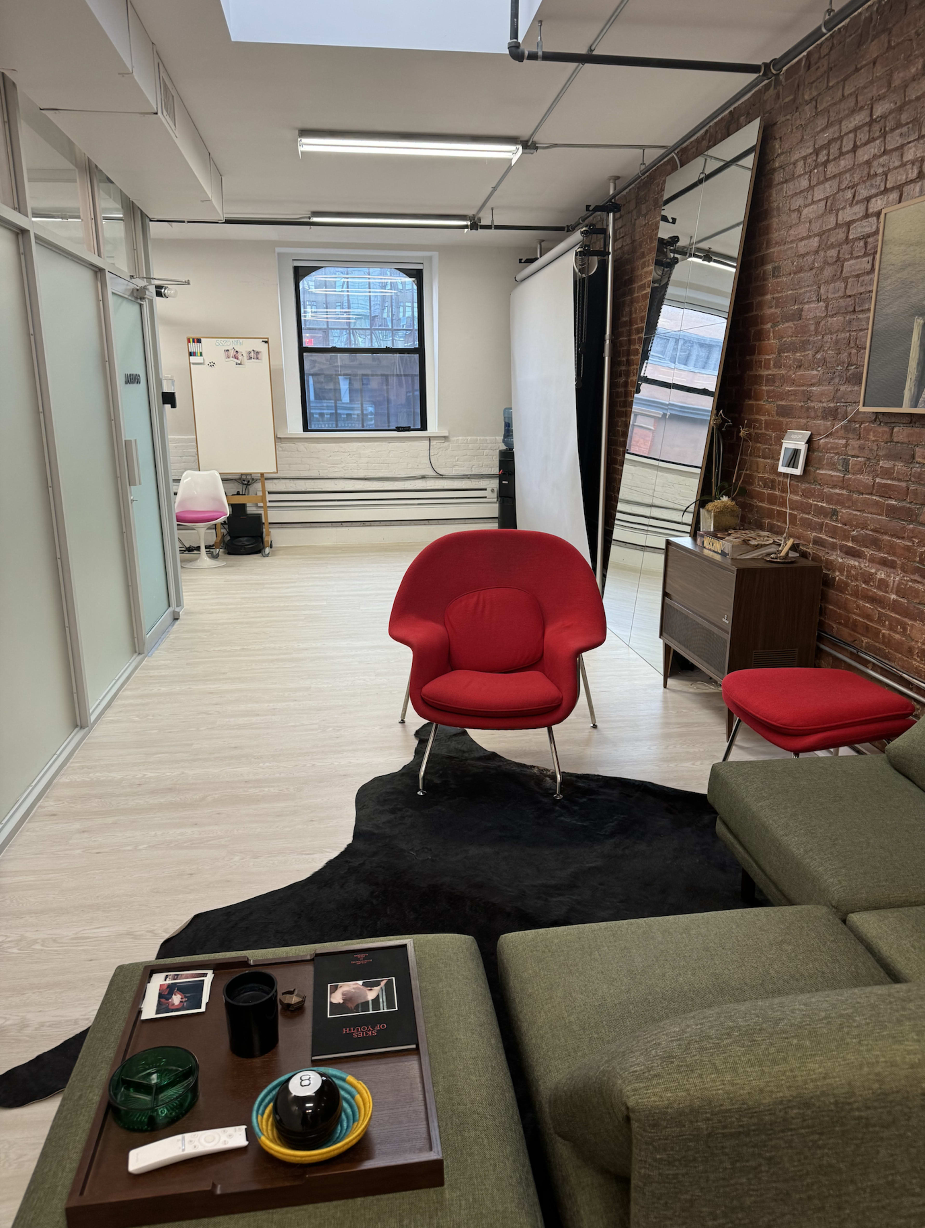 The image shows a modern studio space with a red chair, a green couch, a coffee table, and brick walls, featuring a large mirror and bright lighting.
