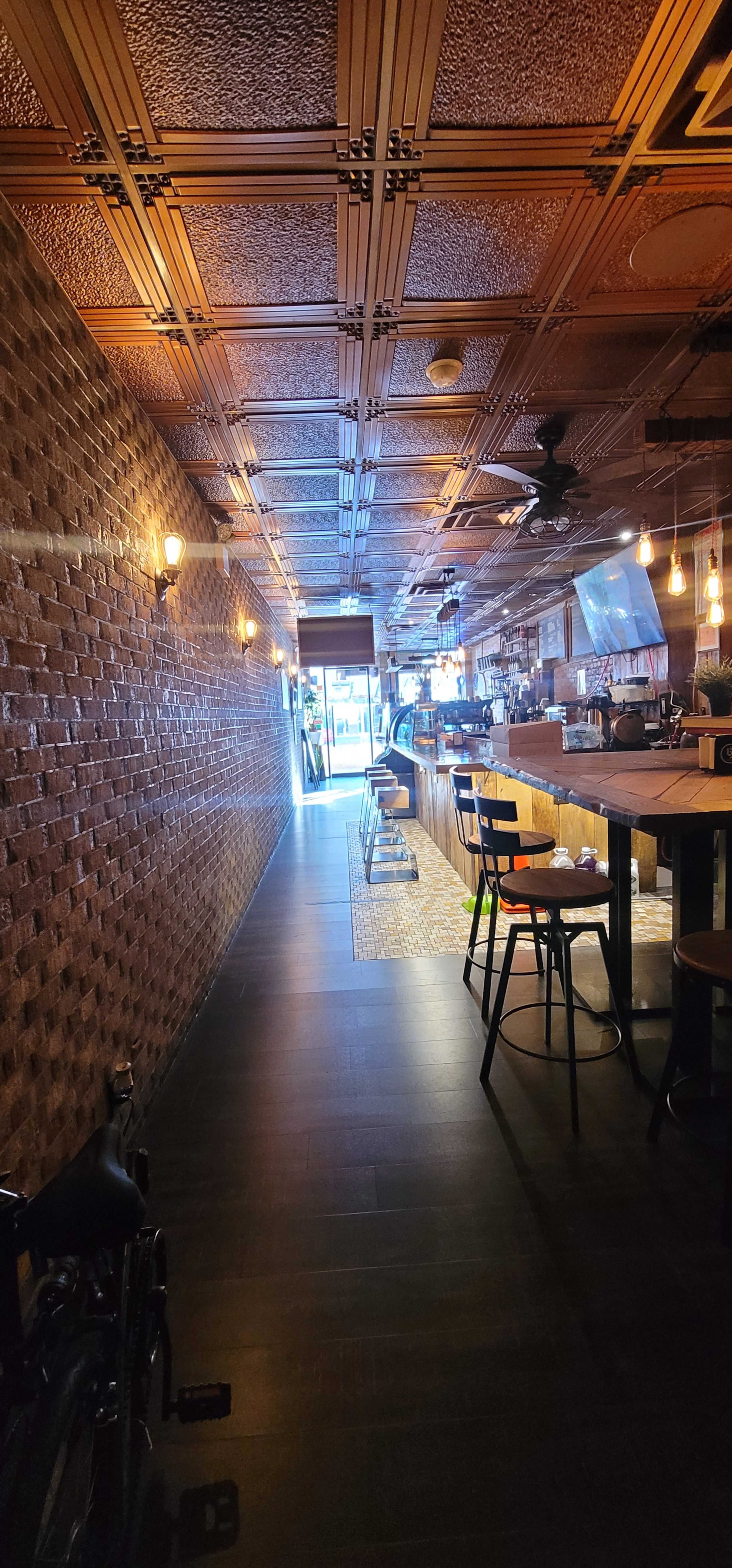 A narrow restaurant interior with a tiled floor, exposed brick walls, and a long bar with high chairs, leading to an entrance illuminated by natural light.