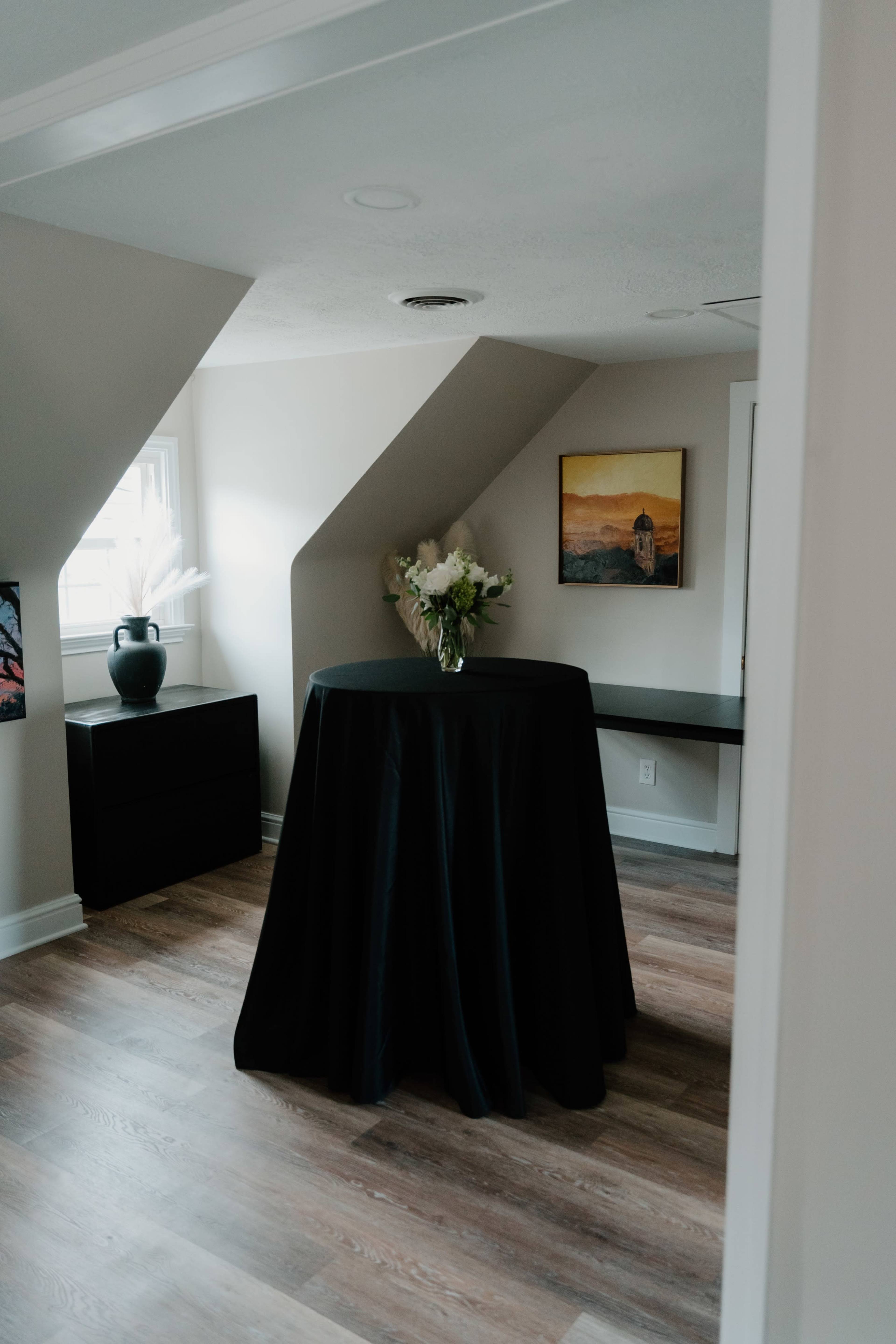 A black table draped with a cloth stands in a corner of a room with angled ceilings, alongside a vase of flowers and a framed painting on the wall.