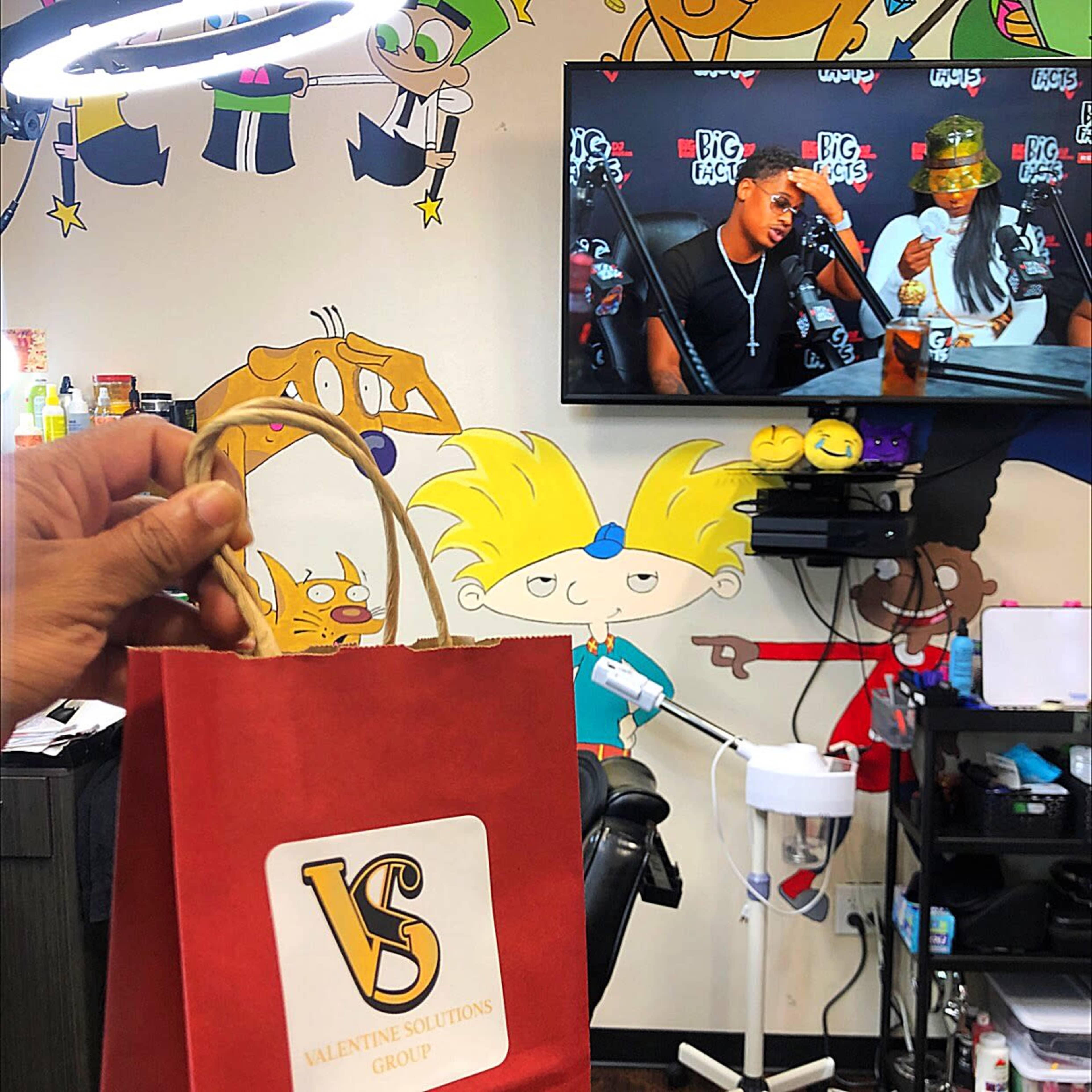 A hand holds a red bag labeled "Valentine Solutions Group" in front of a television displaying a show in a barbershop decorated with cartoon characters on the wall.