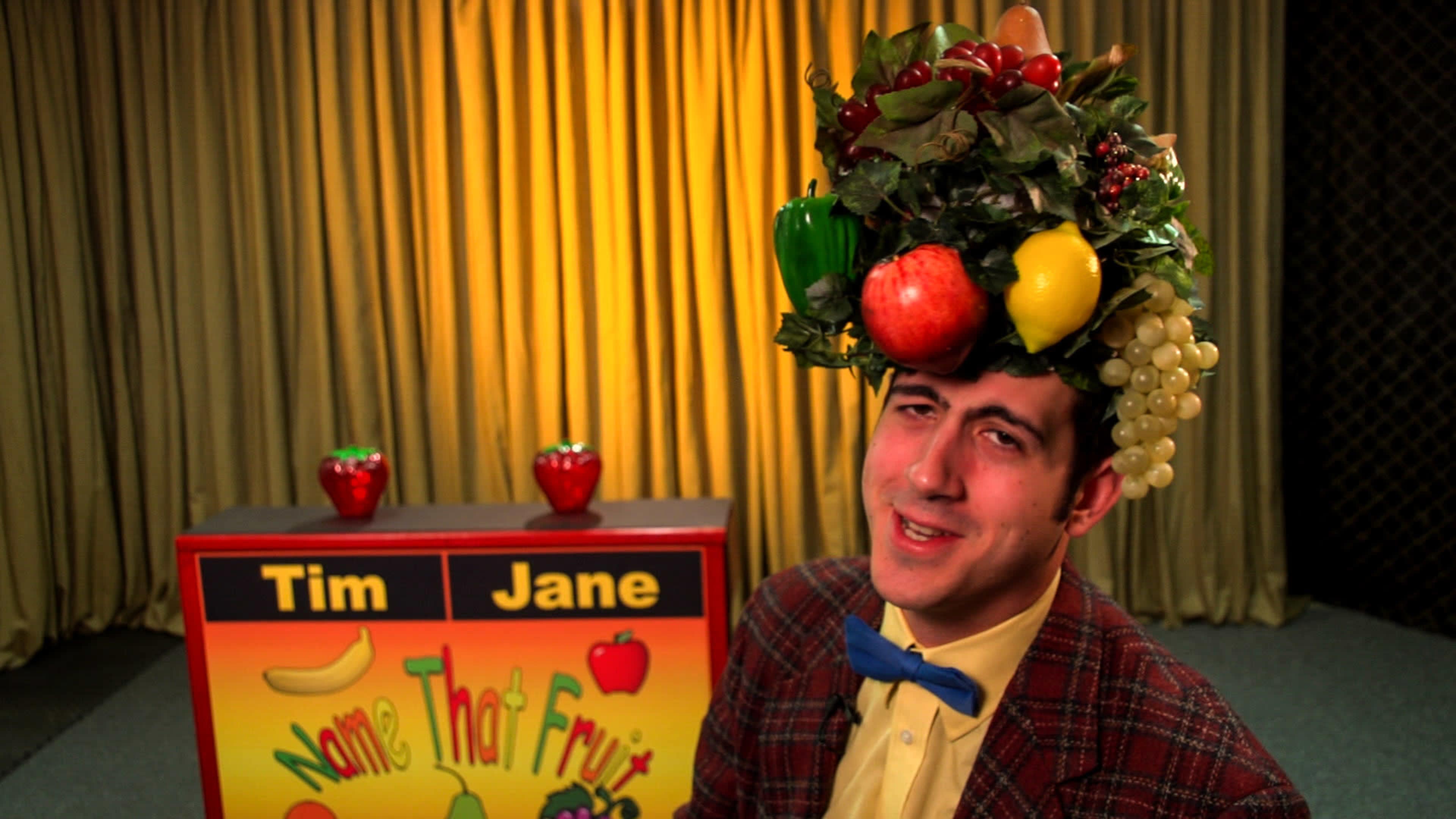 A man wearing a fruit-themed costume stands in front of a game show backdrop labeled "Name That Fruit."