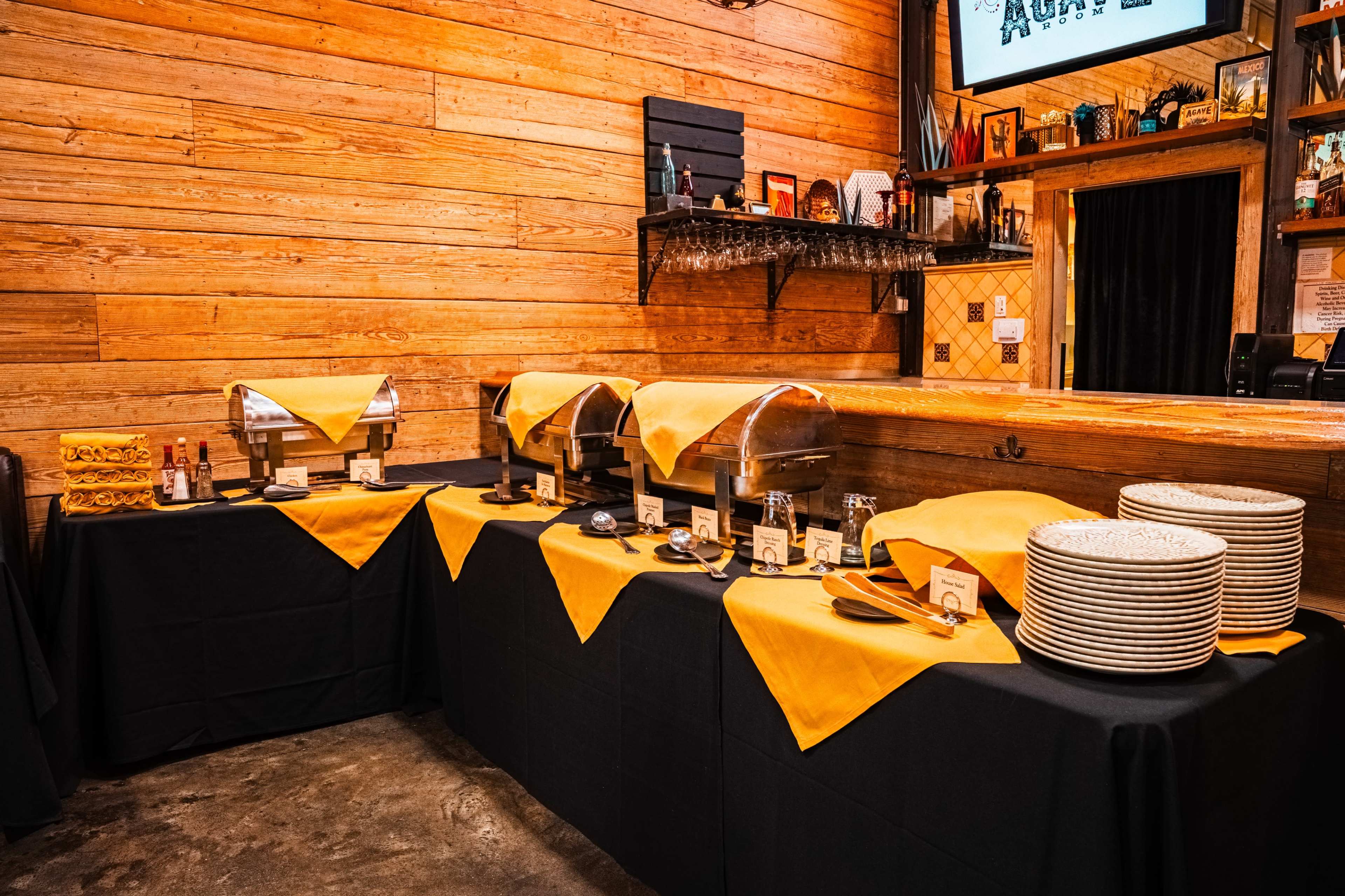 A catering setup displays three chafing dishes covered with yellow cloths on a black table, alongside stacks of plates and condiment bottles against a wooden wall.