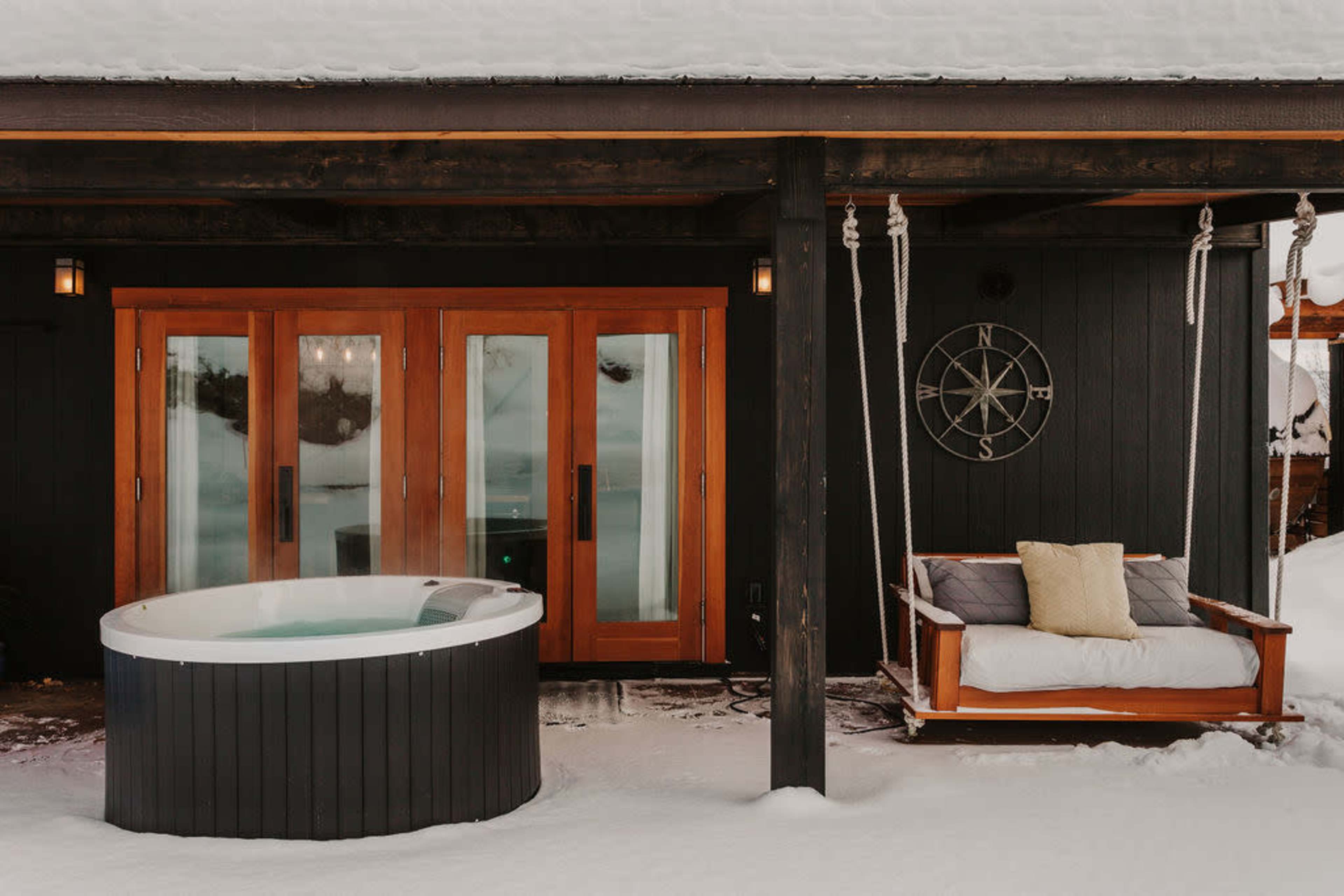 A snowy outdoor space featuring a hot tub and a swing bench beside a dark wooden structure with large glass doors.