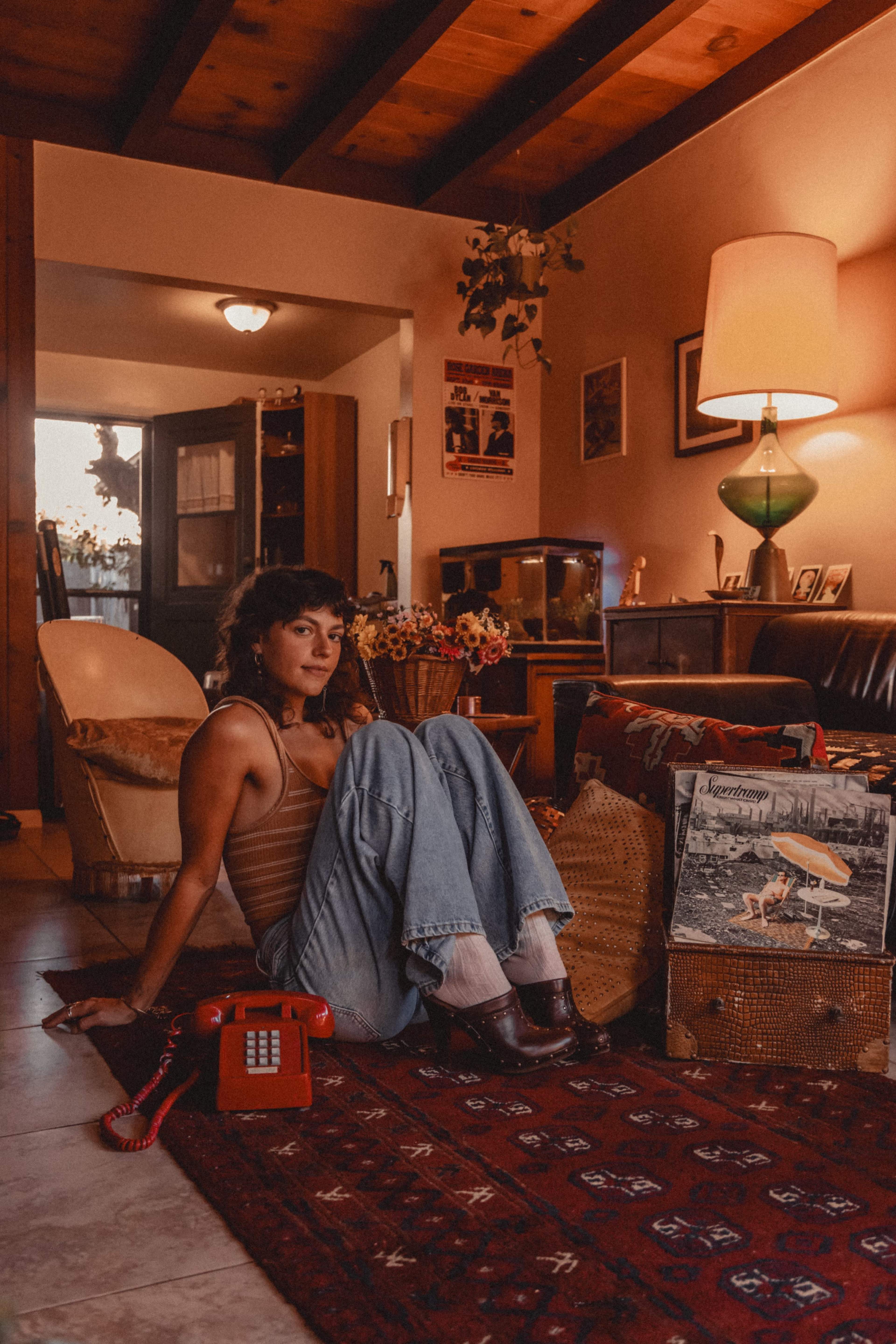 A woman sits on the floor in a cozy living room, surrounded by vintage decor, including a red telephone and a stack of records.