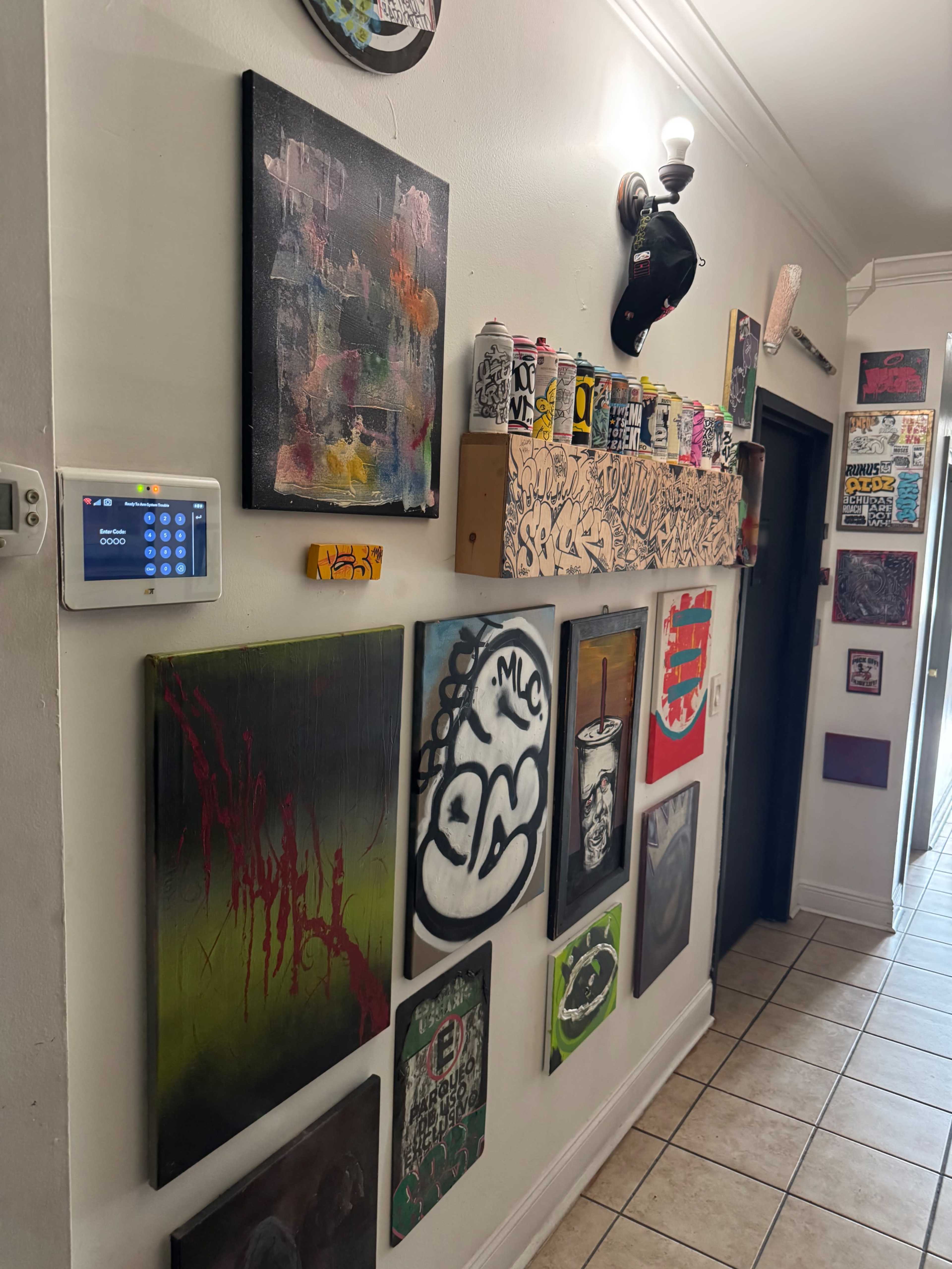 The image shows a hallway adorned with various pieces of artwork, including graffiti-style paintings and a shelf displaying spray paint cans and other art supplies.