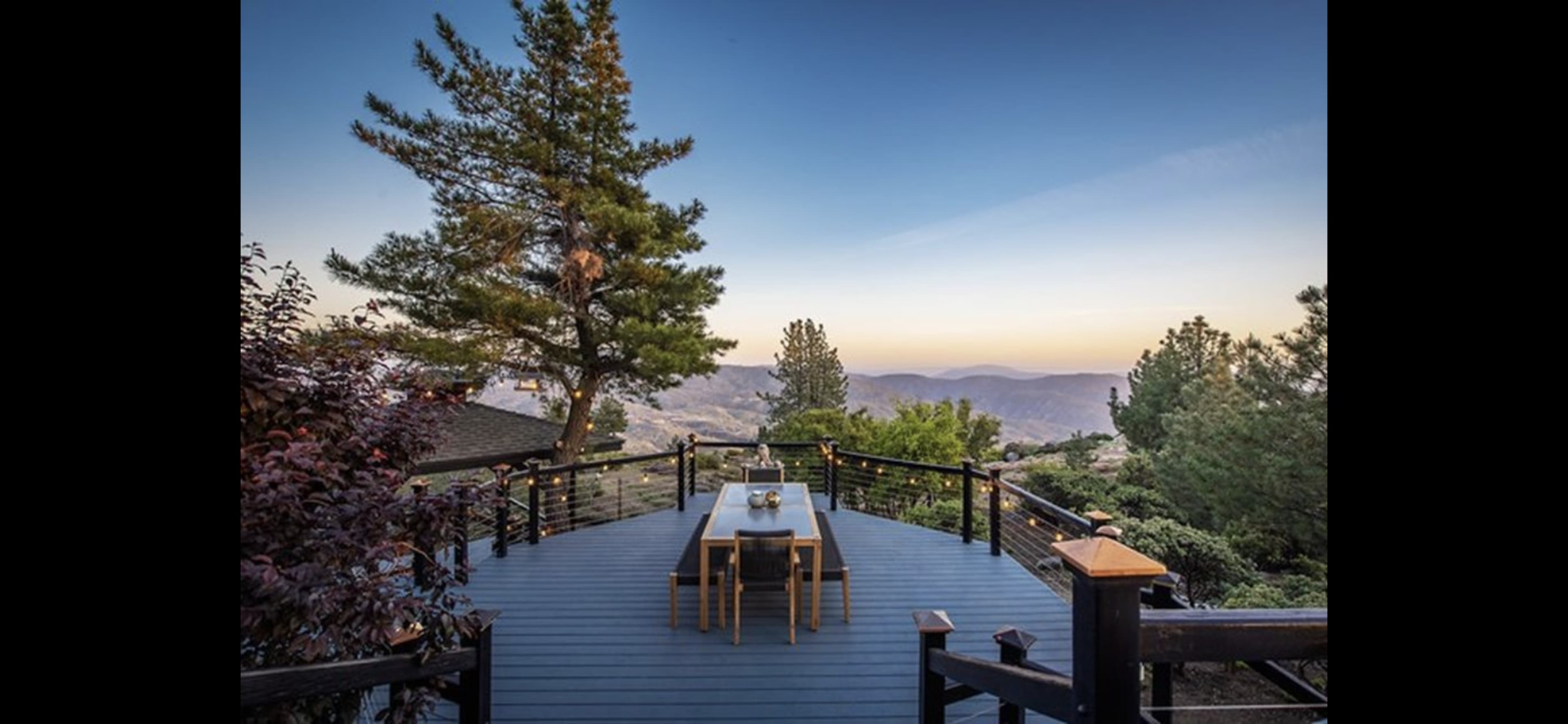 A wooden deck with a long table overlooks a mountainous landscape at dusk, surrounded by trees.