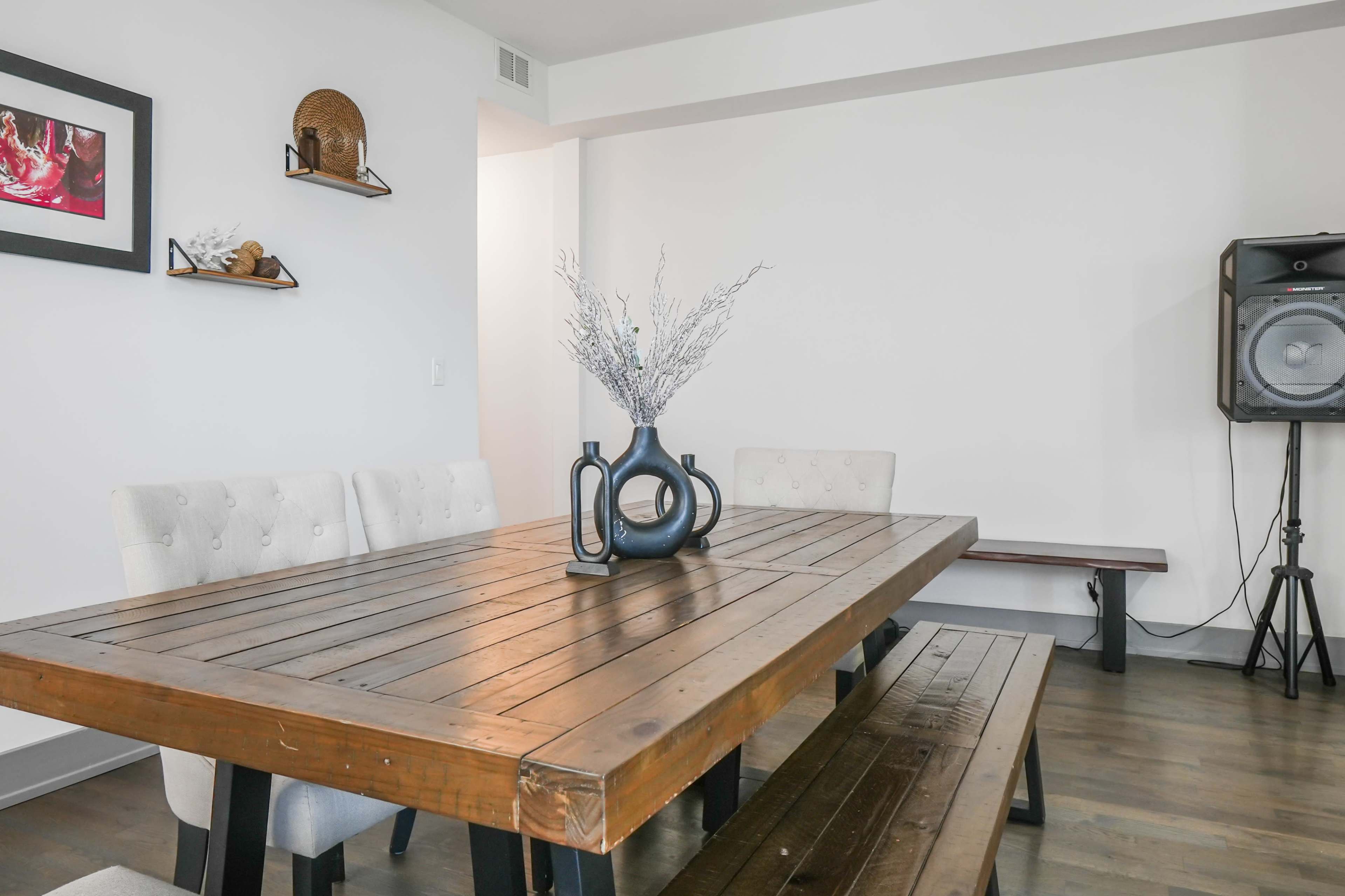 A wooden dining table with benches is set in a well-lit room, featuring a vase with flowers at its center and speaker equipment in the background.