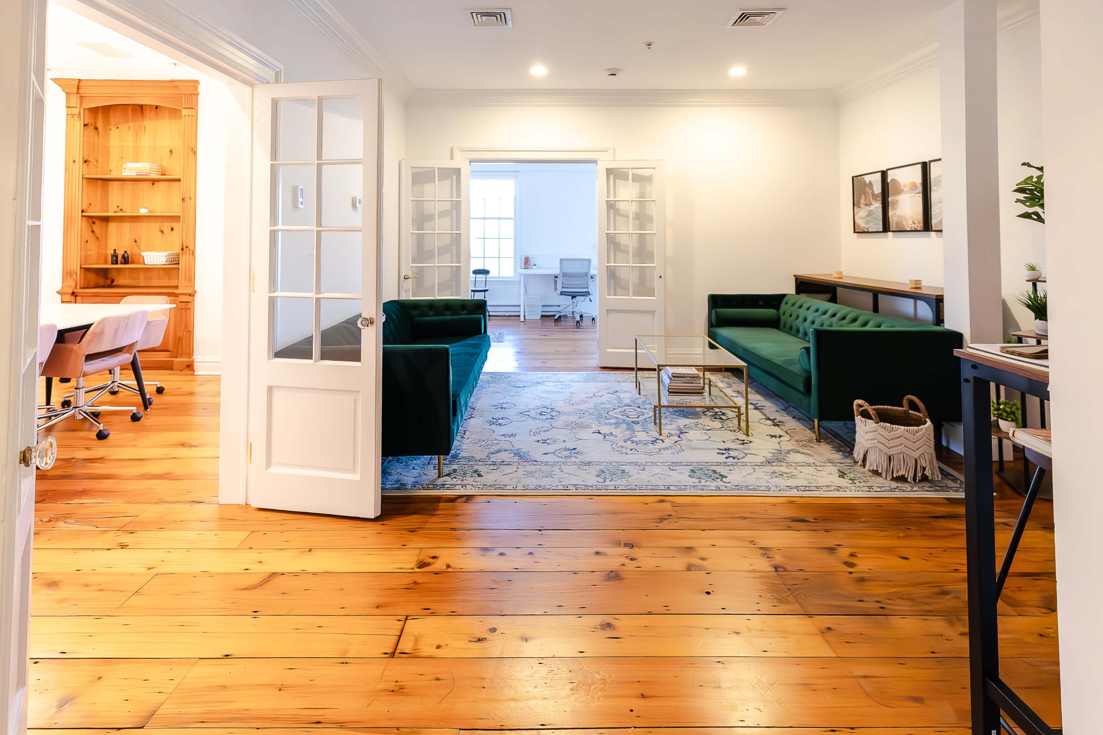 A well-lit office space featuring green sofas, a coffee table, and wooden flooring, with an adjacent room visible through open double doors.