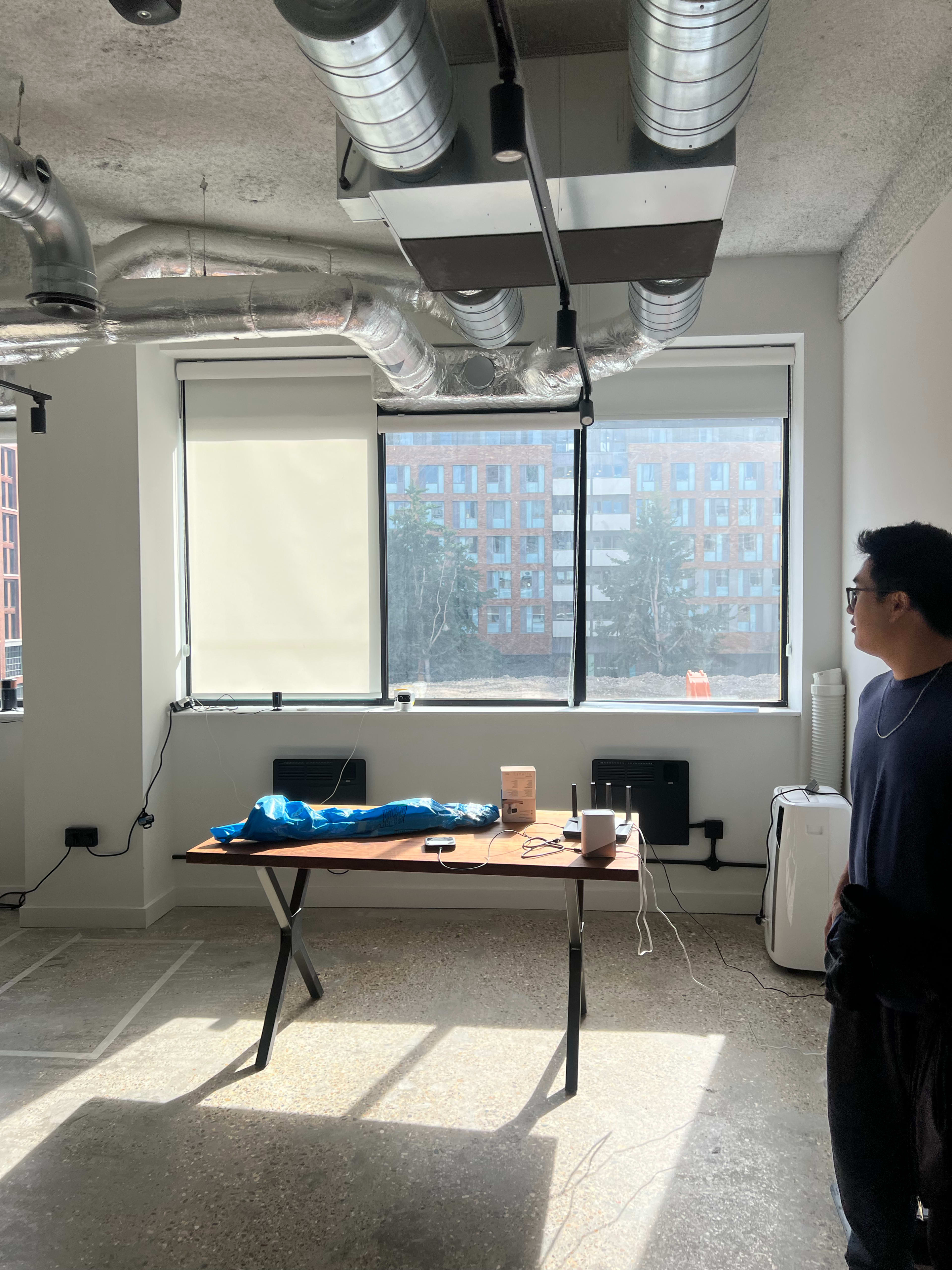 The image shows a modern office space with exposed ductwork, a table featuring various items, and a large window overlooking a building complex.
