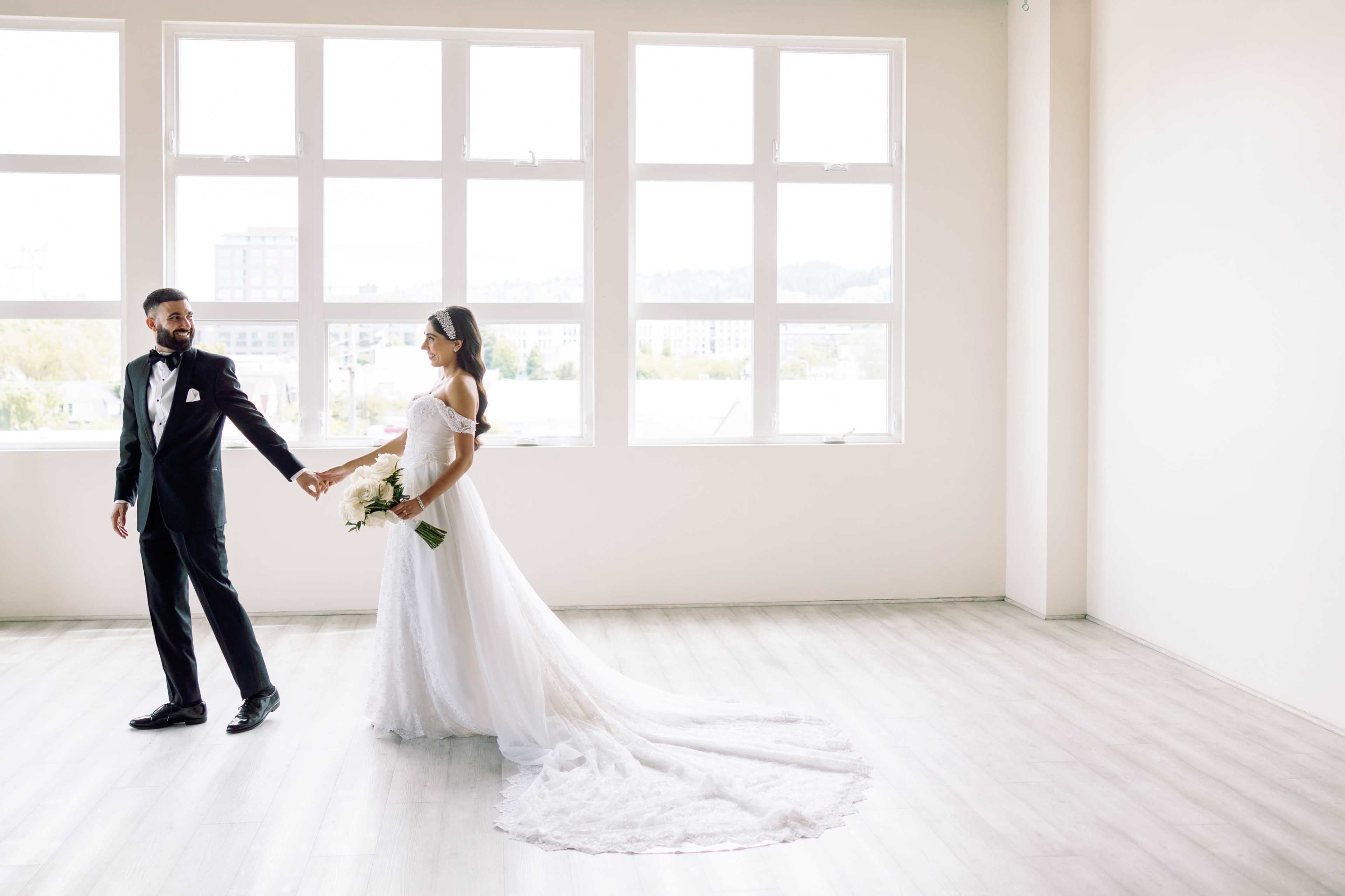 A bride and groom hold hands and walk together in a spacious, well-lit room with large windows.
