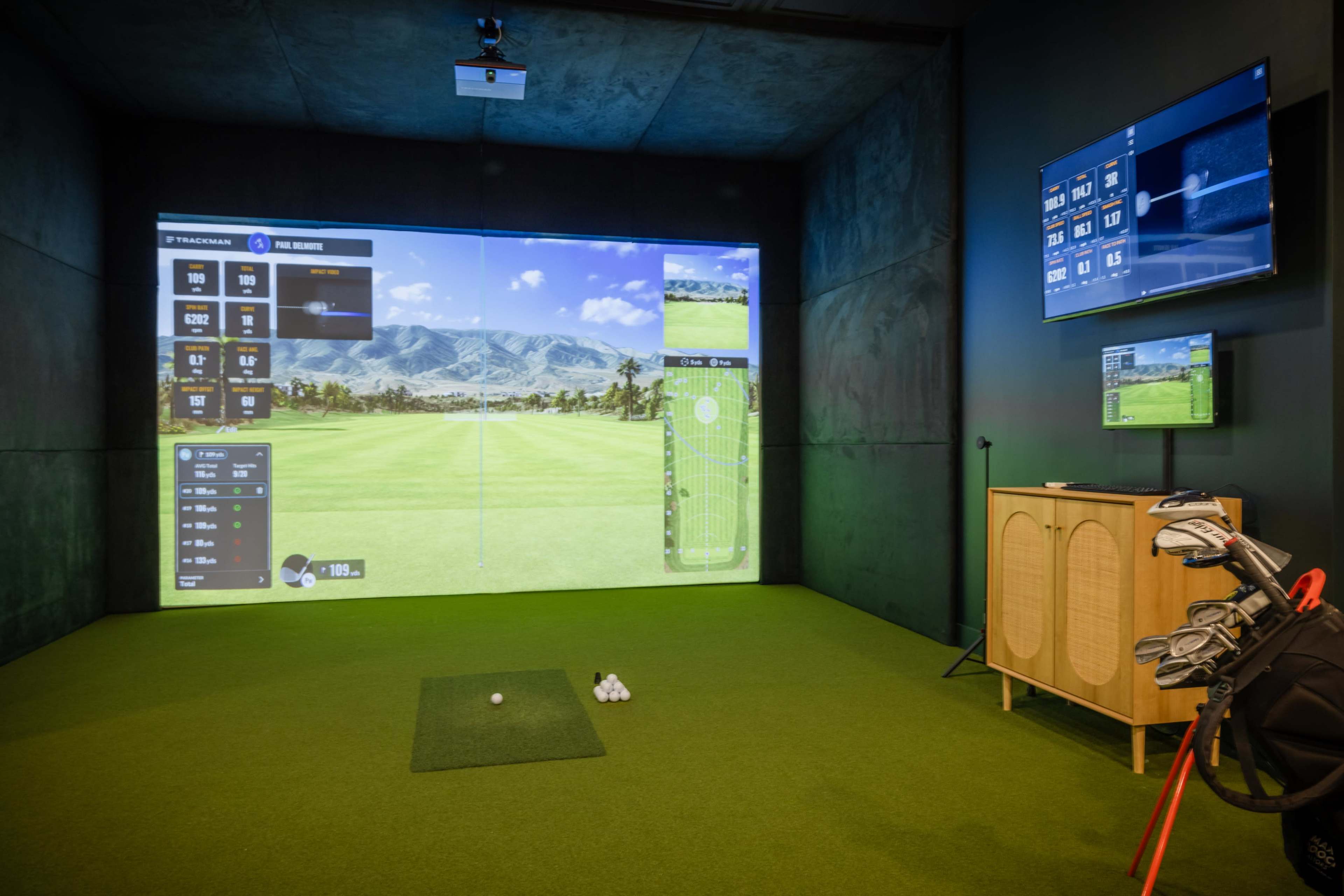 An indoor golf simulator setup with a large projection screen displaying a golf course, a golf tee with balls, and a set of clubs beside a cabinet.