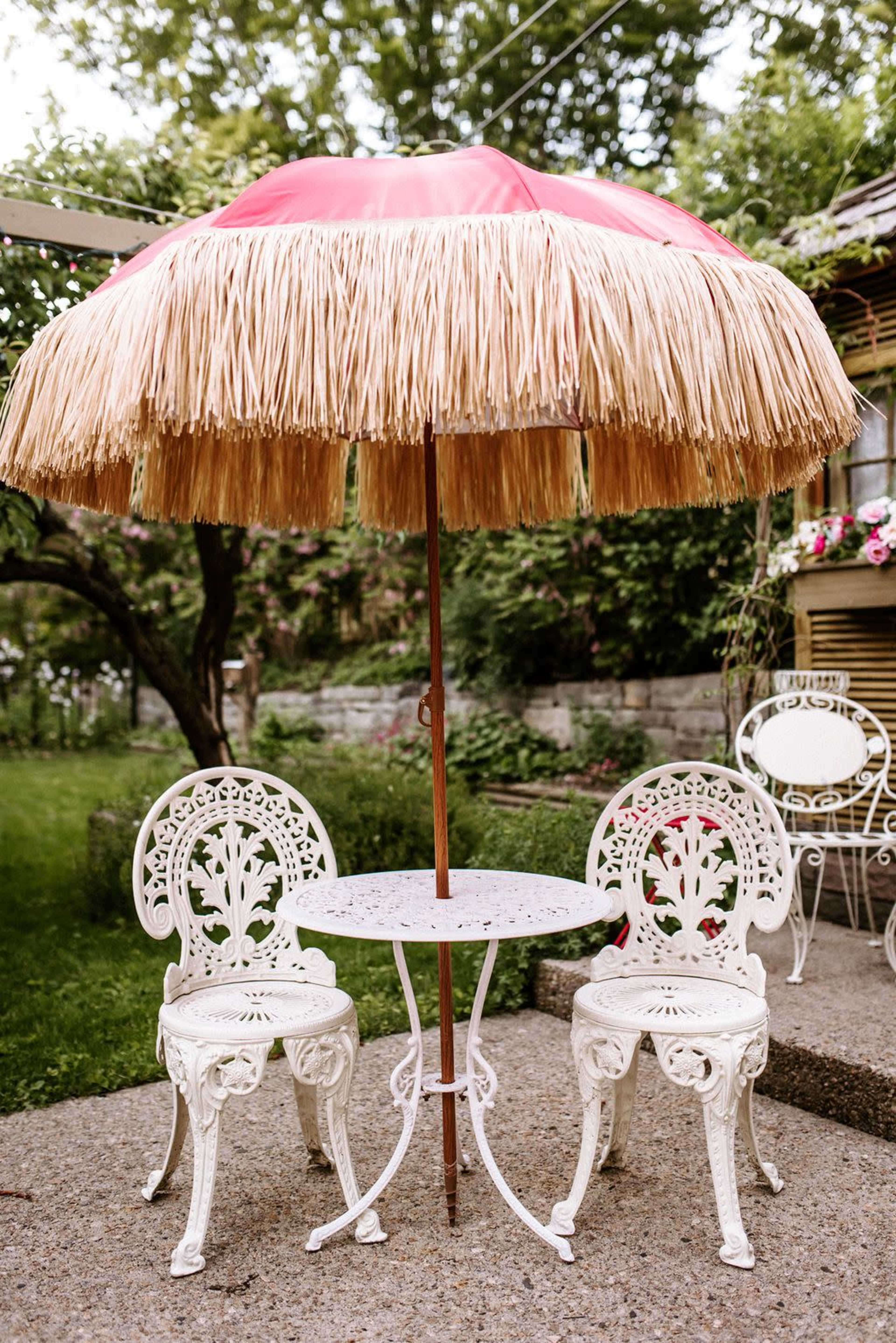 A round table with two white wrought iron chairs is situated under a large, pink thatched umbrella in a garden setting.