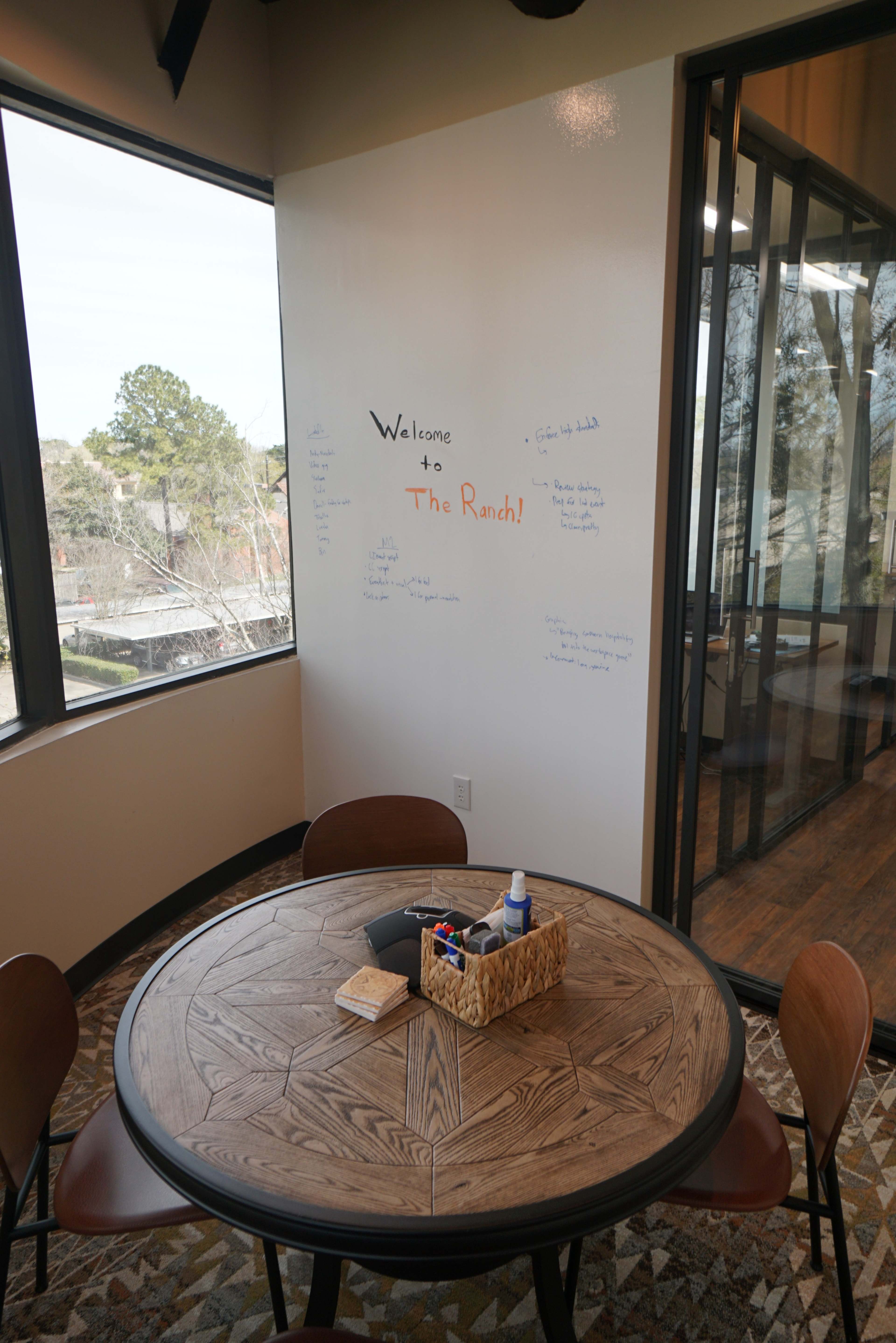 A round wooden table with chairs is situated in a bright room featuring a whiteboard that says "Welcome to The Ranch."