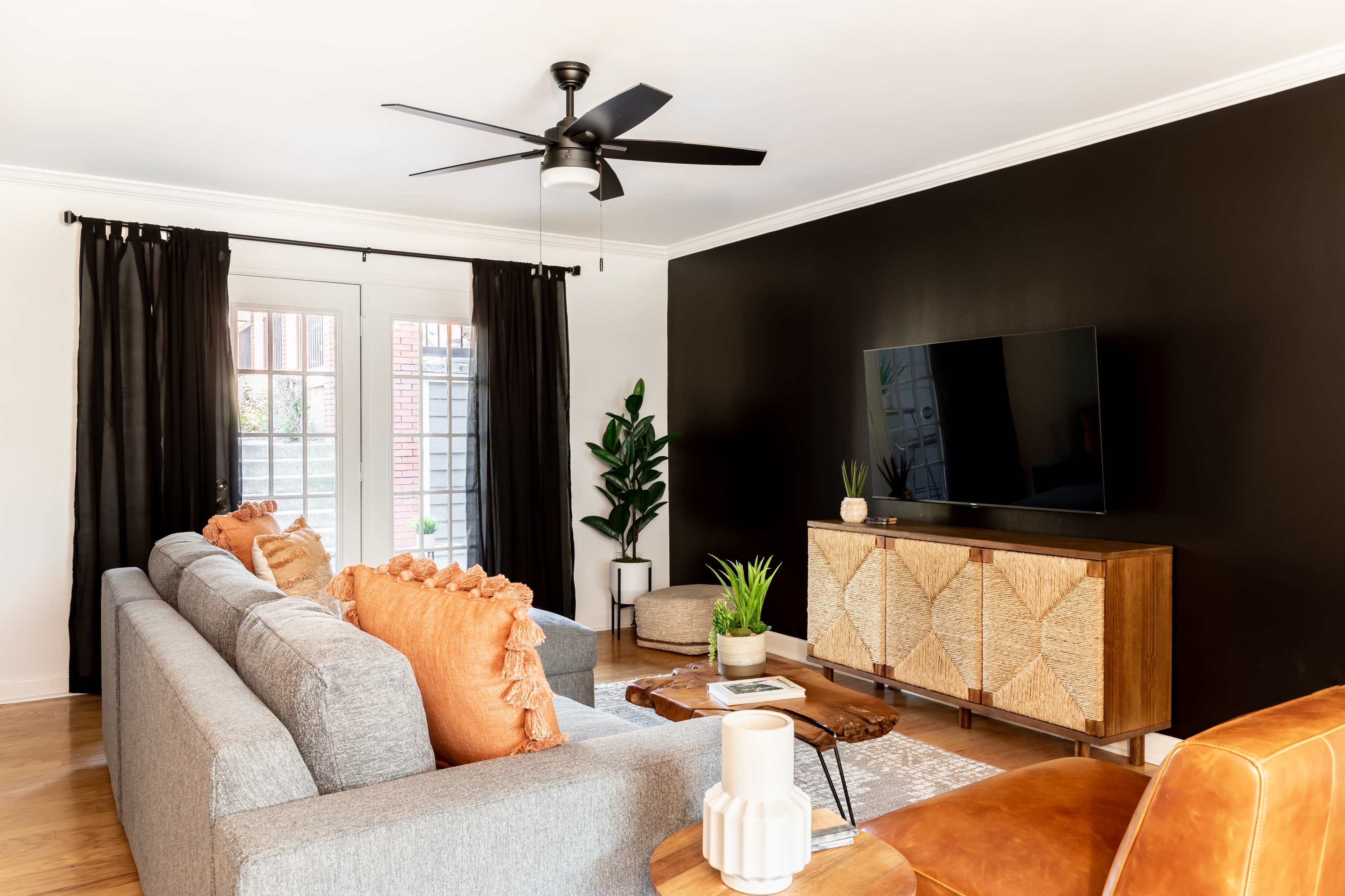 A modern living room features a gray sofa with orange cushions, a wooden media console, a TV mounted on a black wall, and potted plants near a door with black curtains.