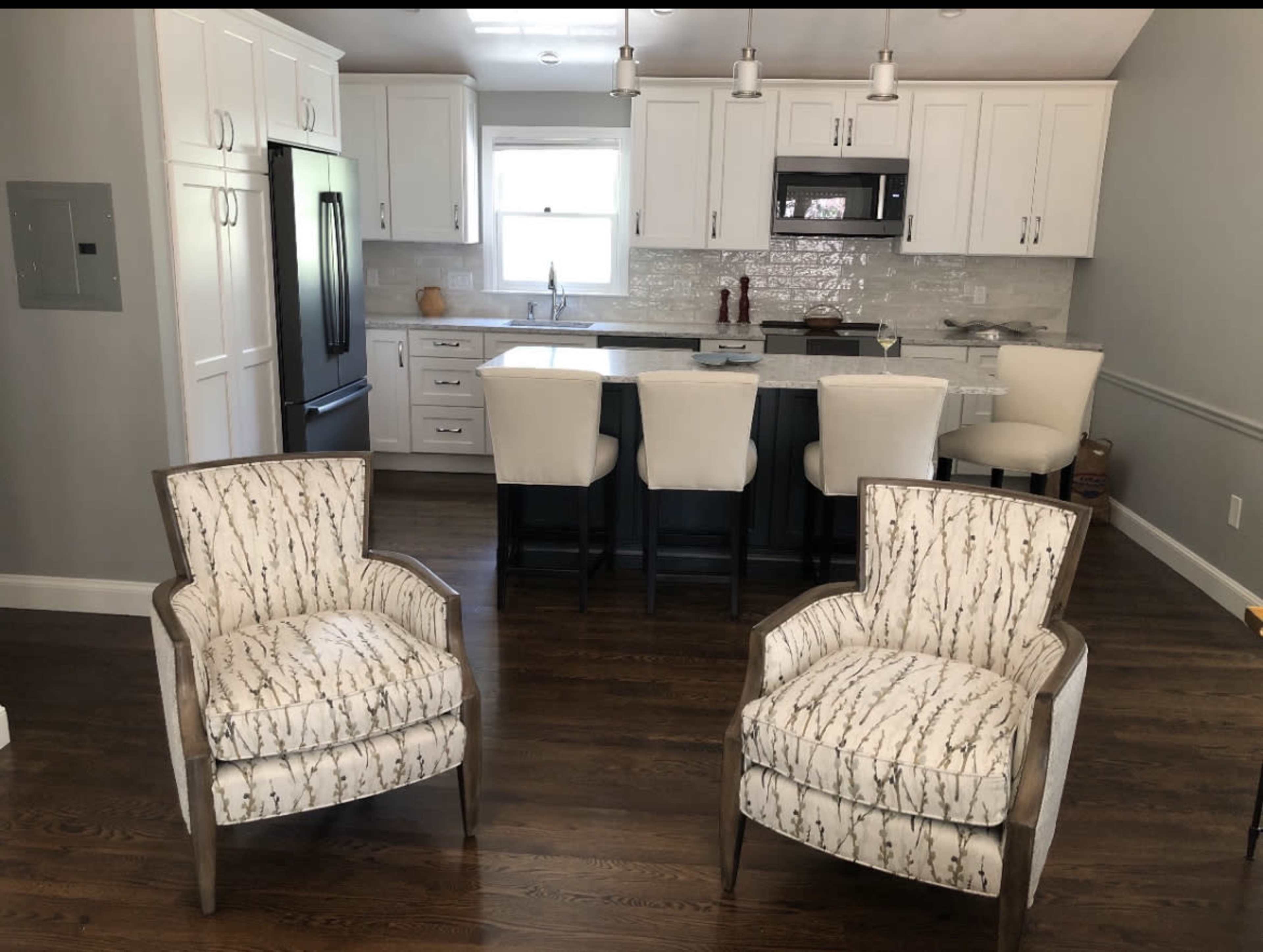 A modern kitchen features a central island with four bar stools, surrounded by white cabinetry and stainless steel appliances, with two patterned chairs positioned in the foreground.