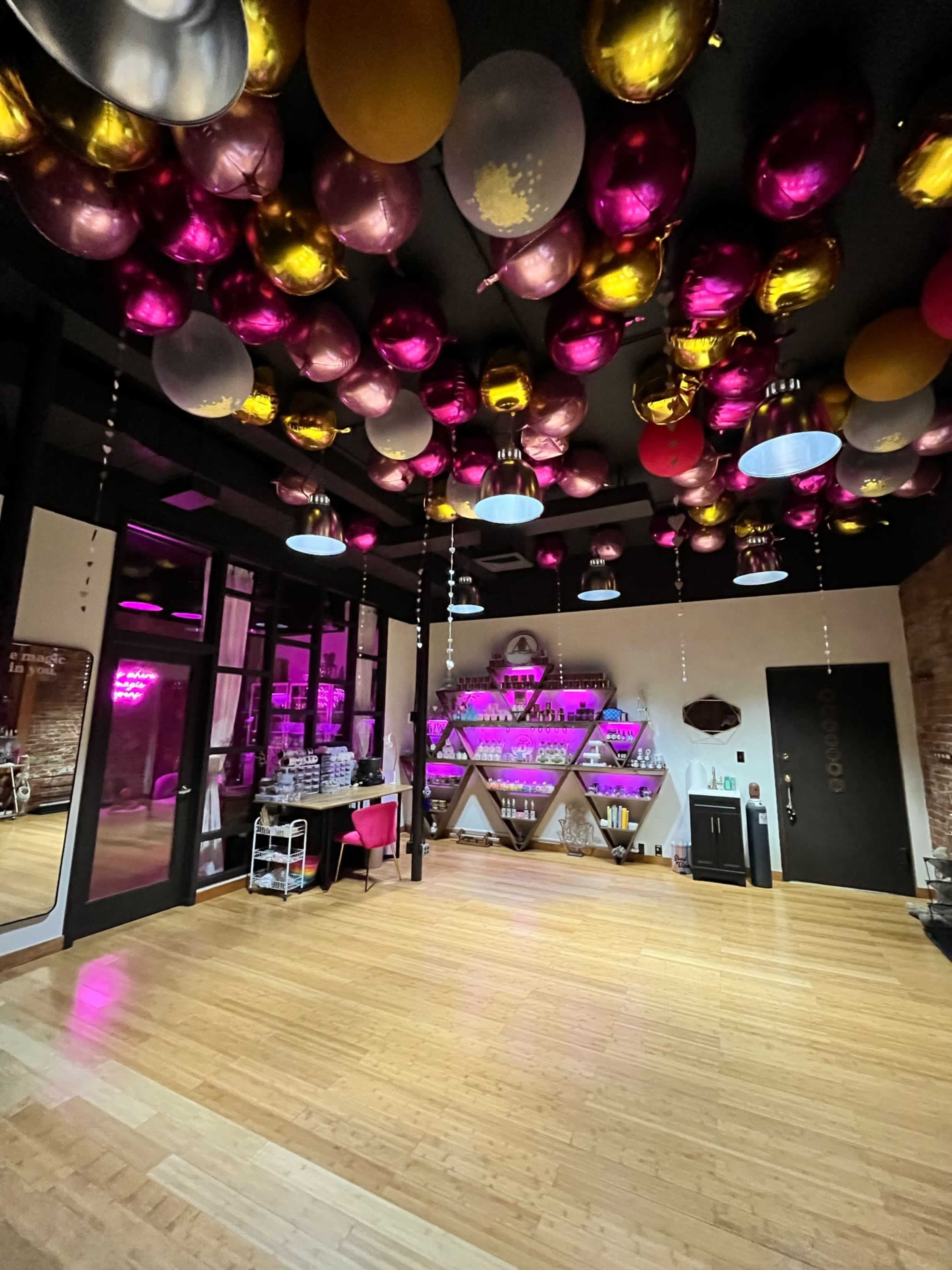 The interior of a venue features a wooden floor, a wall adorned with shelves of colorful bottles, and a ceiling decorated with pink and gold balloons.