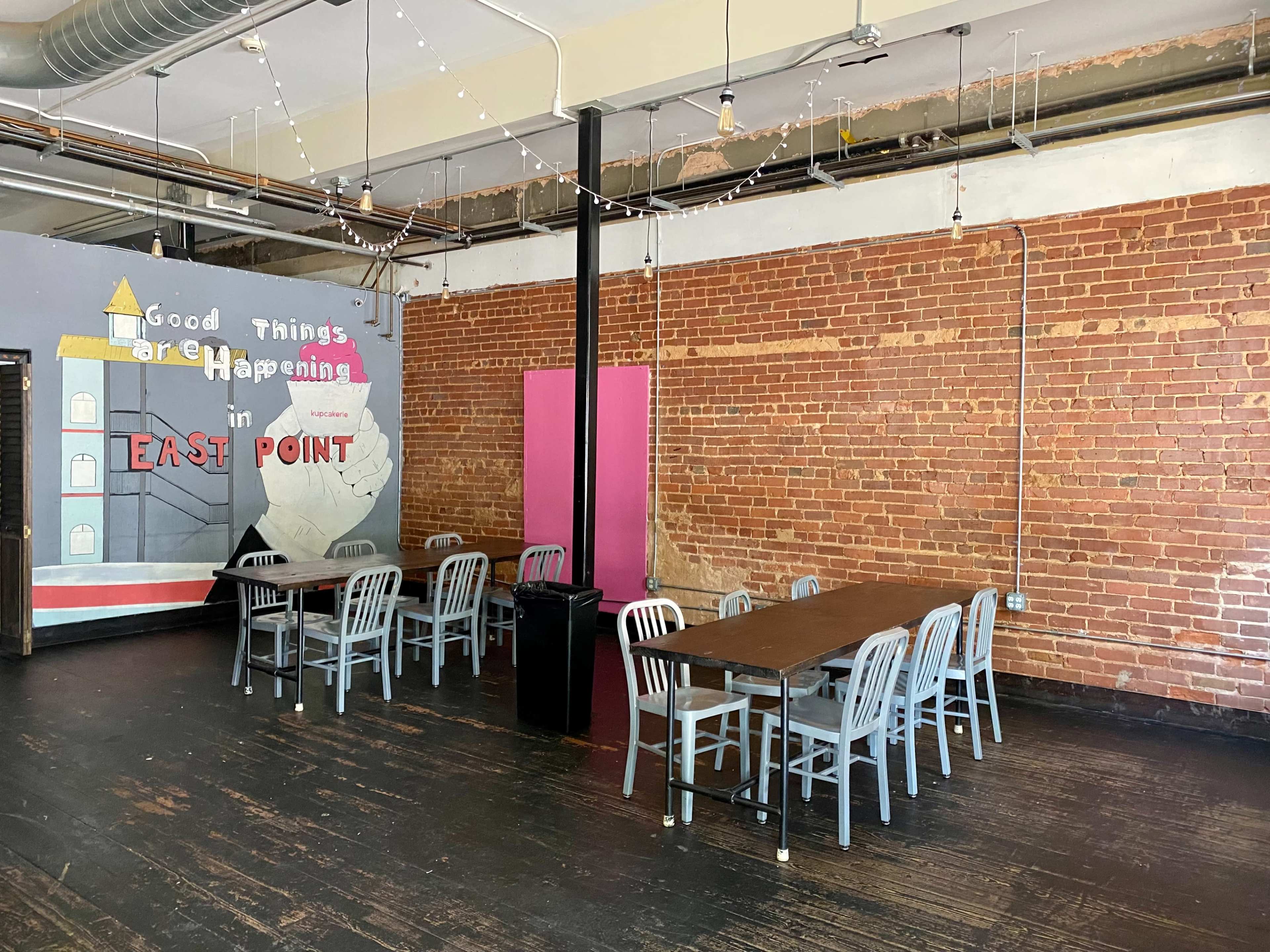 A spacious room with a long table surrounded by chairs, exposed brick walls, and a mural that reads "Good Things Are Happening in East Point."