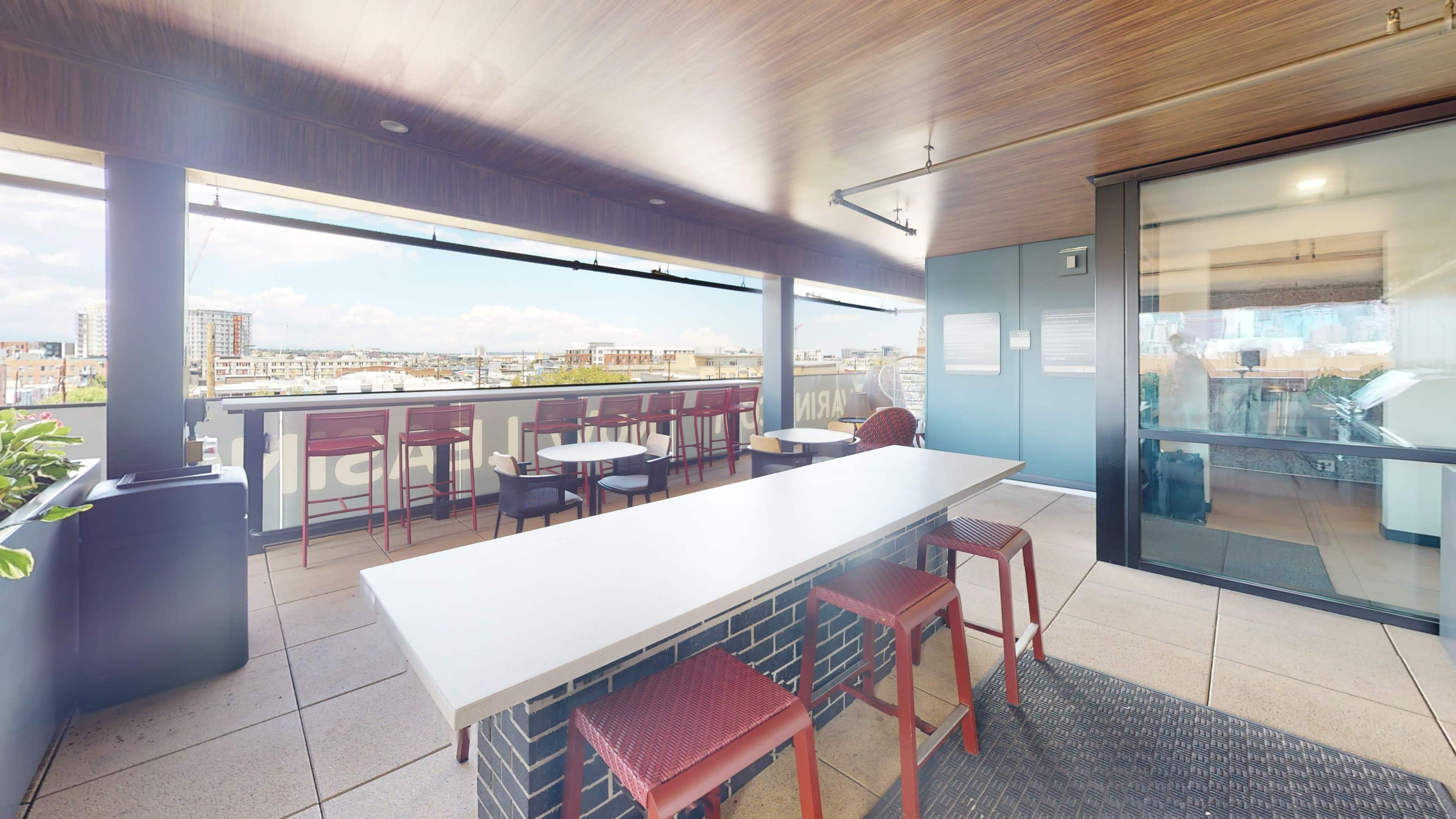 The image shows a rooftop terrace with a large table and red chairs, overlooking a cityscape with buildings and a clear sky.
