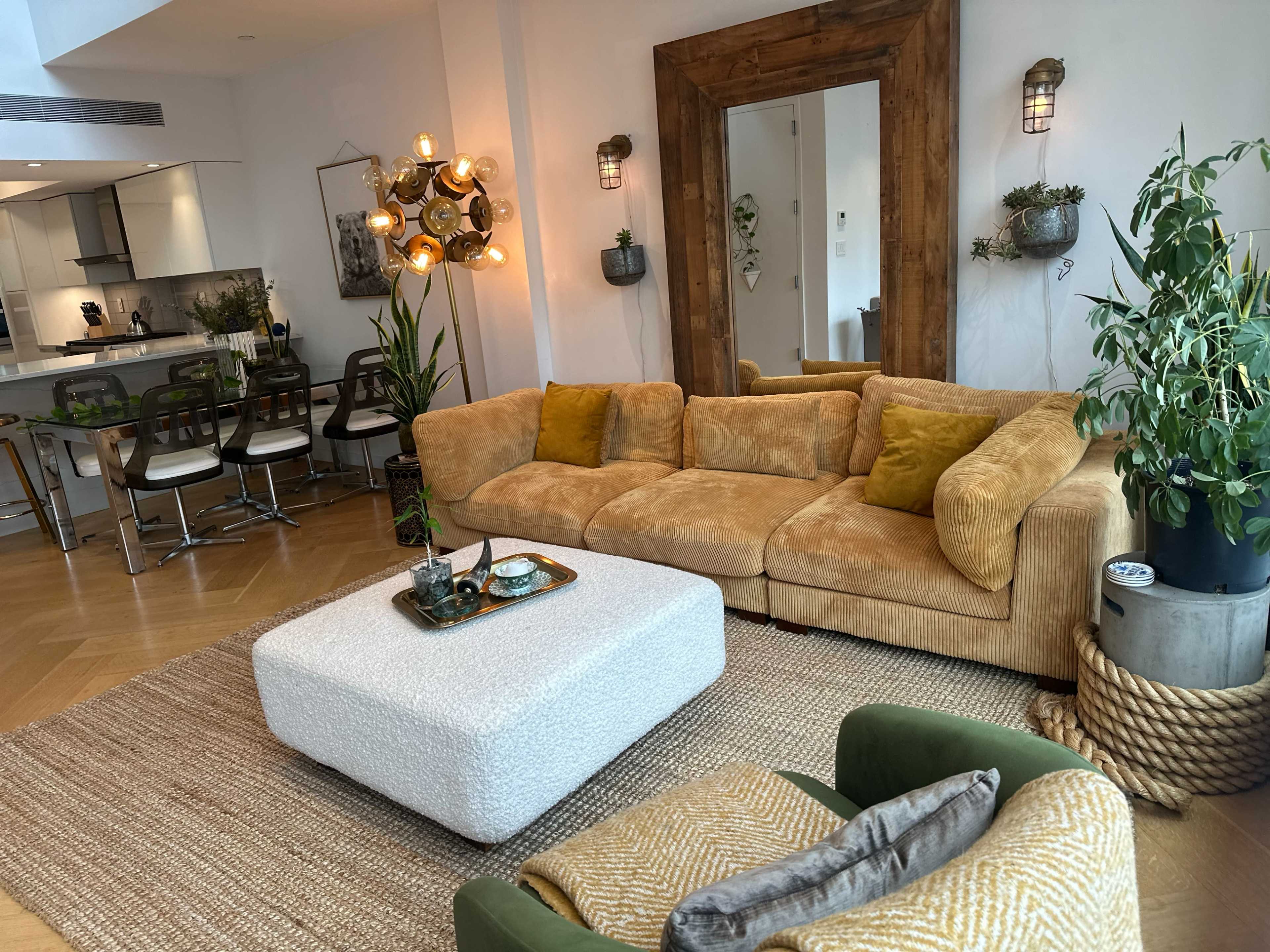 The image shows a modern living room featuring a large, mustard-colored sectional sofa, a light-colored ottoman, and a mirror framed in wood, with plants and decorative items integrated into the decor.