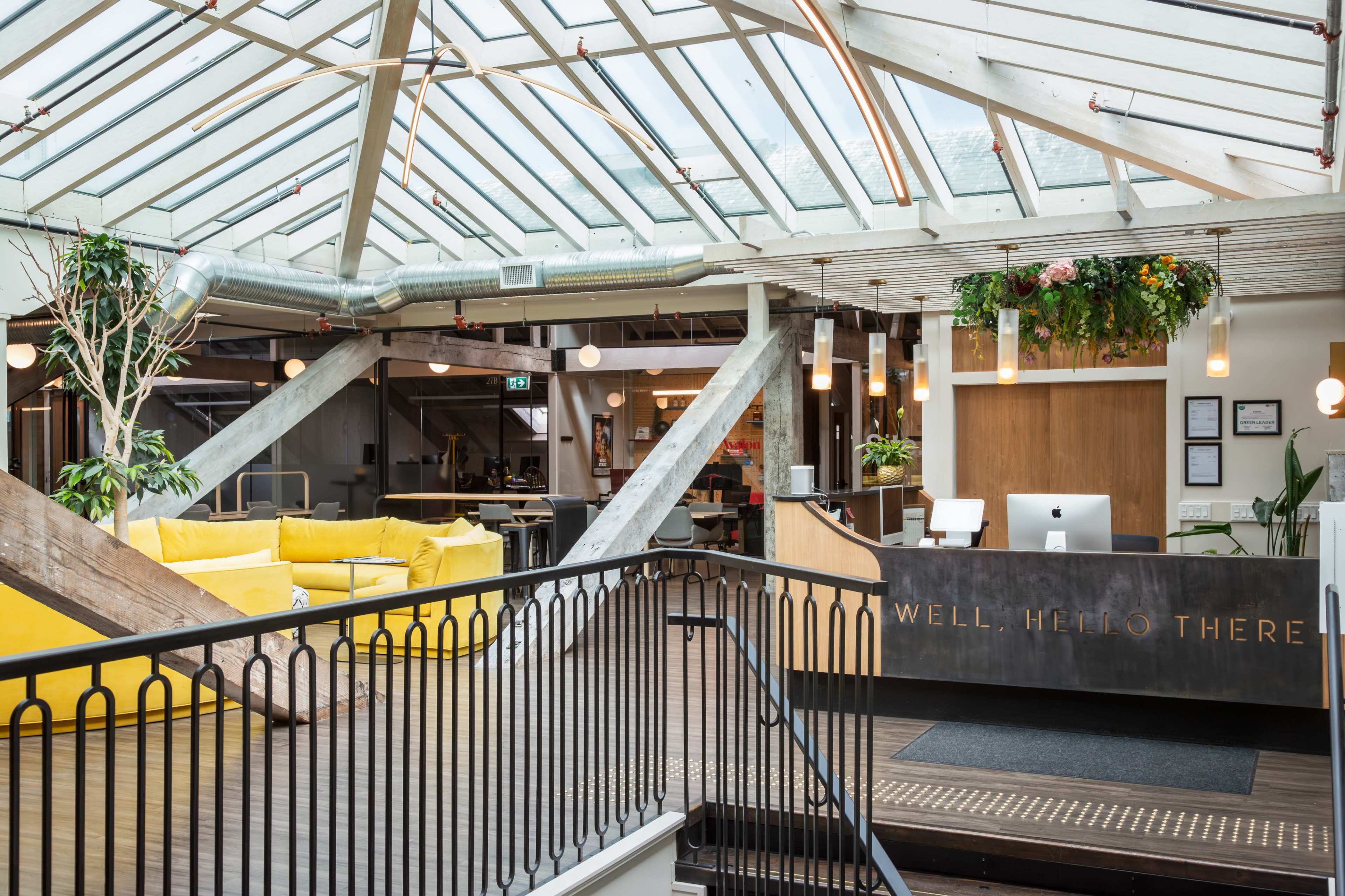 The image shows a modern, open-concept office space with a reception area and a large skylight, featuring wooden beams and vibrant yellow seating.