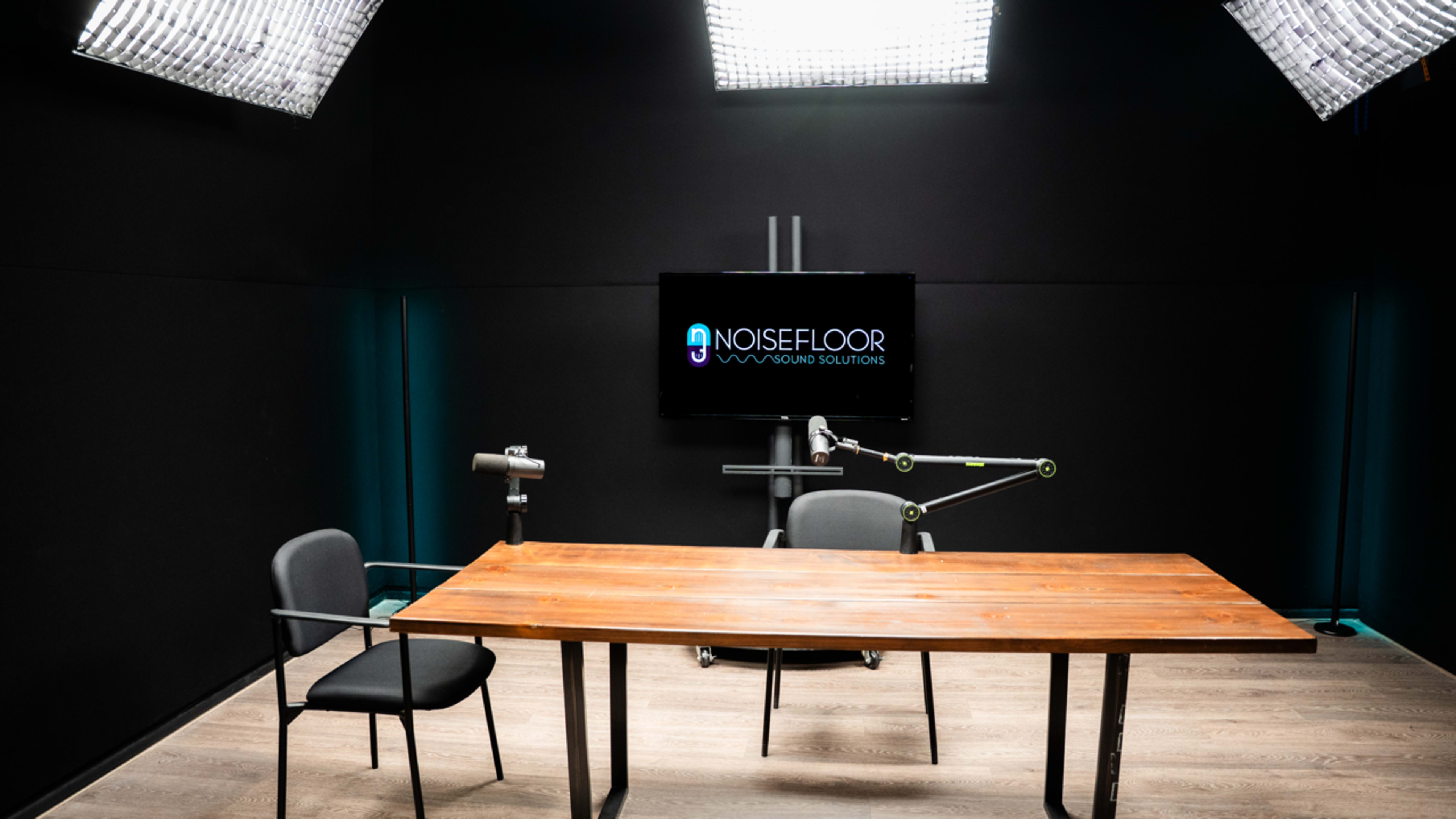 A studio setup features a wooden table and one chair, with lighting panels and a screen displaying the logo "NOISEFLOOR" on a black wall.