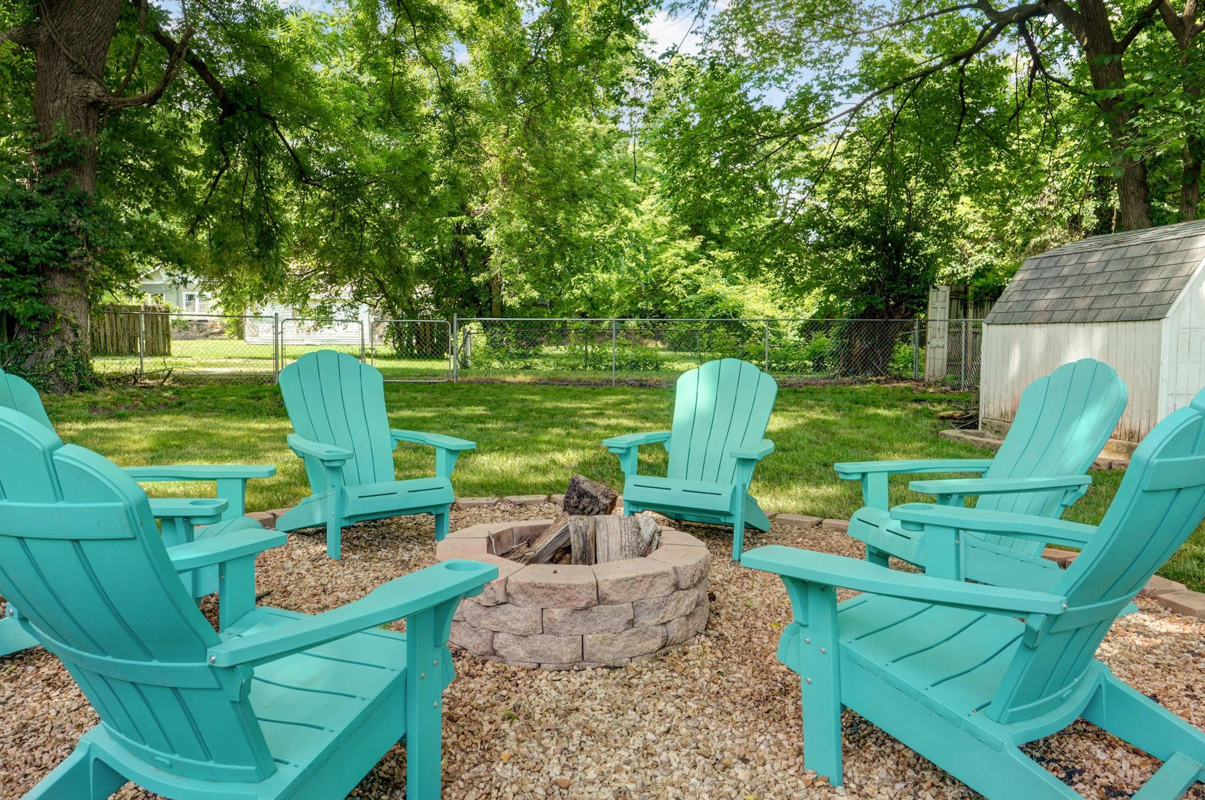 A circular fire pit surrounded by six turquoise Adirondack chairs sits in a grassy backyard with trees and a shed in the background.