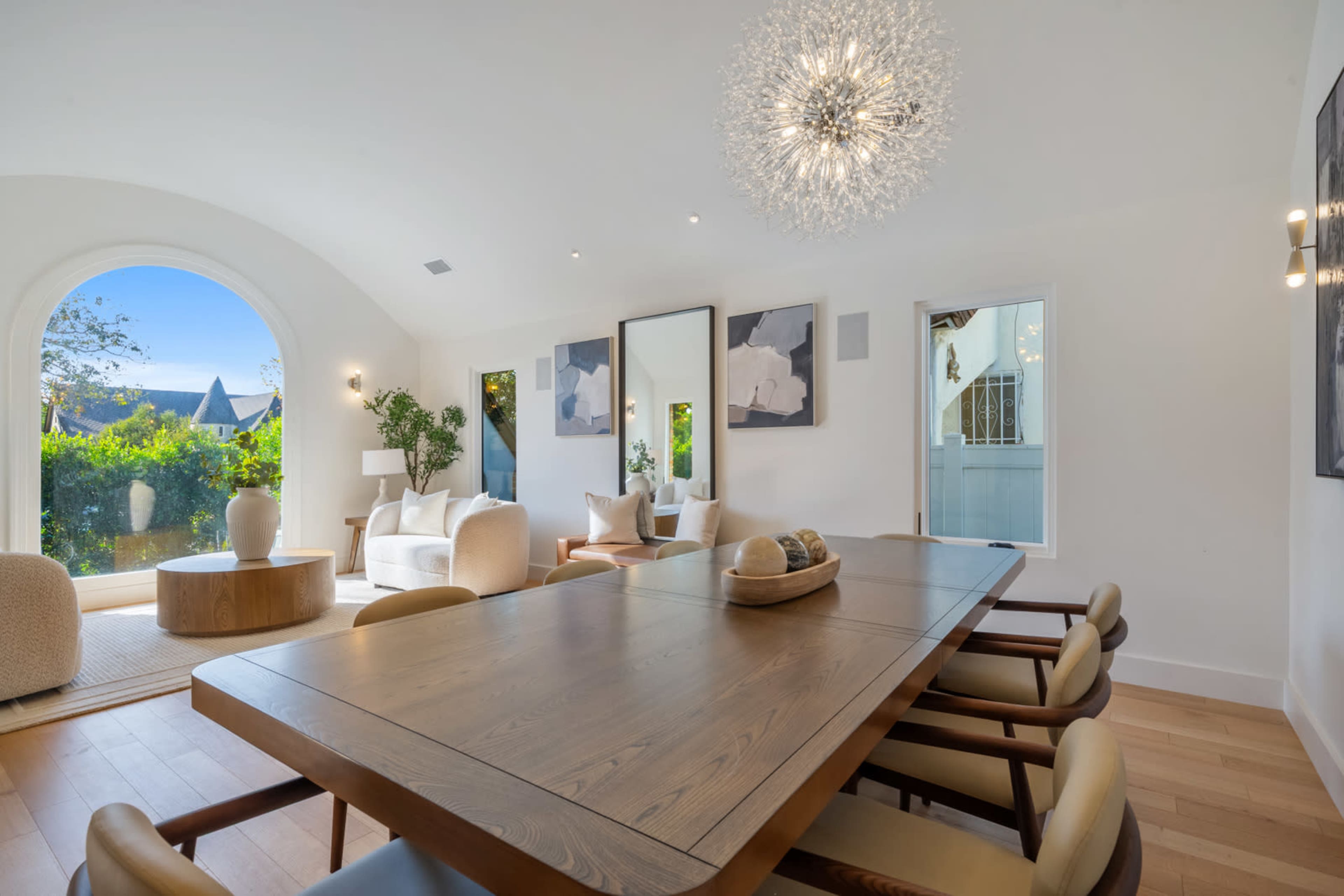 A modern dining area features a wooden table surrounded by beige chairs, with large windows displaying greenery outside.