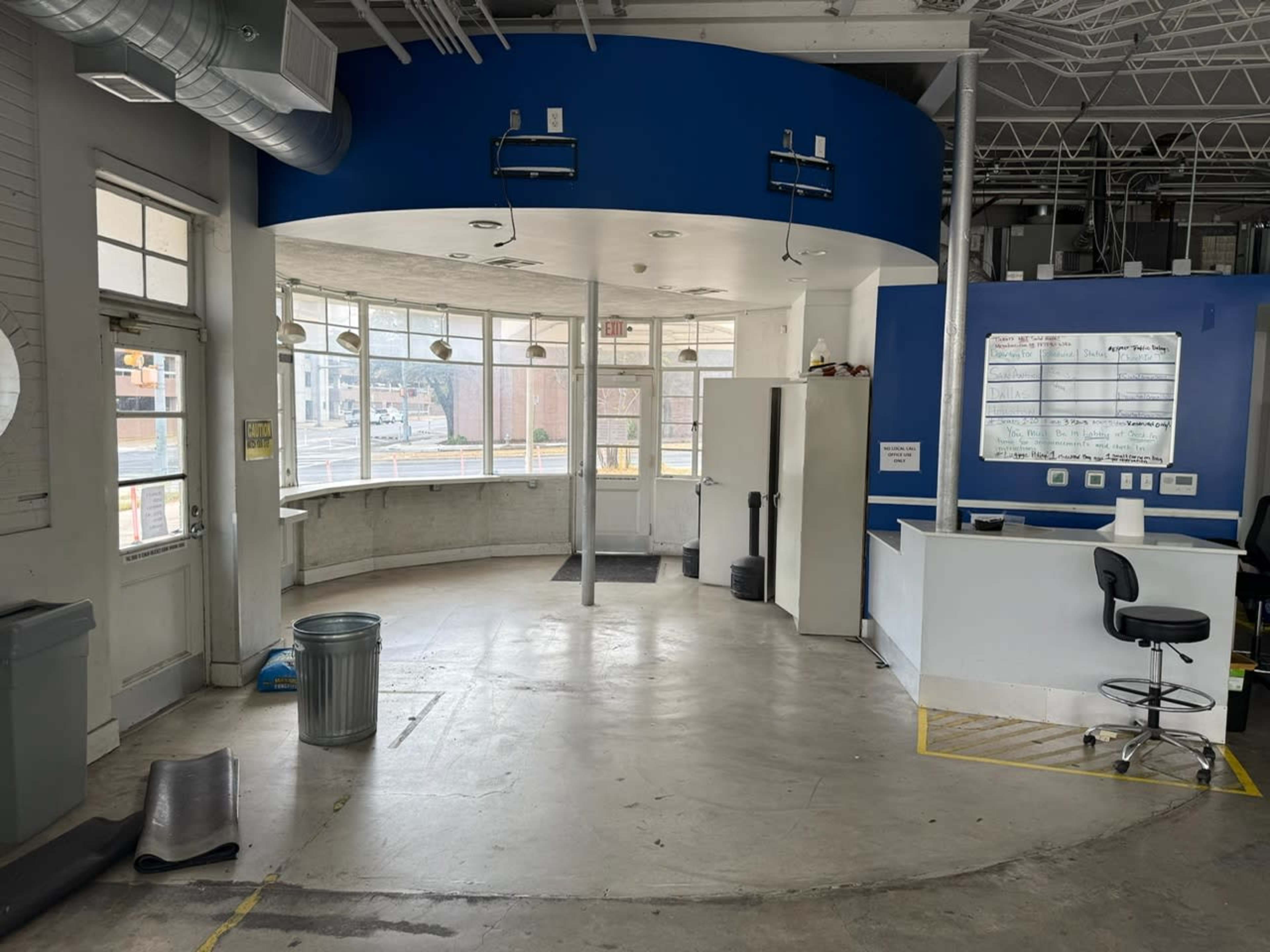 The image shows a spacious reception area with large windows, a front desk, a whiteboard, and a trash can in a modern building.