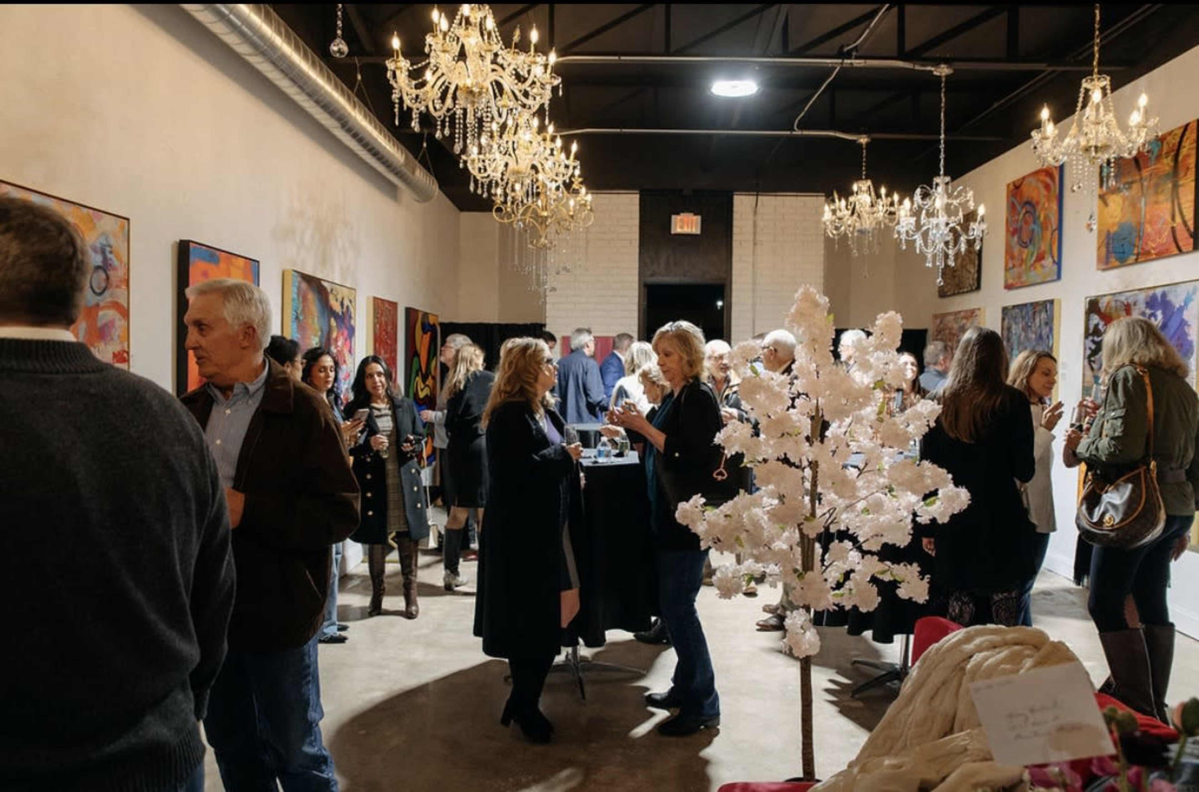 A gallery event features a crowd of people mingling among colorful artwork and decorative chandeliers.