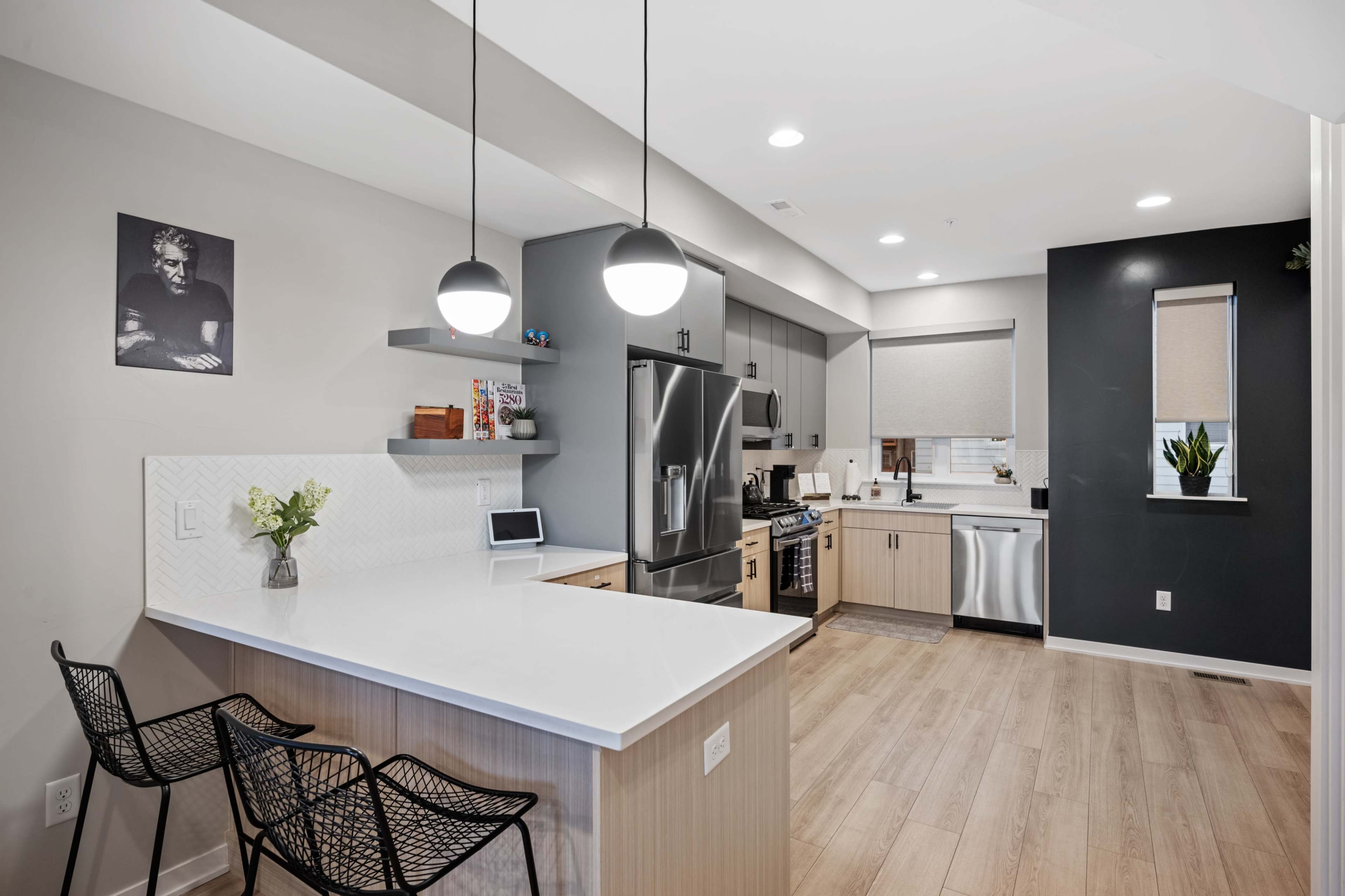 A modern kitchen with a white countertop, stainless steel appliances, and black accents features two bar stools and a wall-mounted photo.