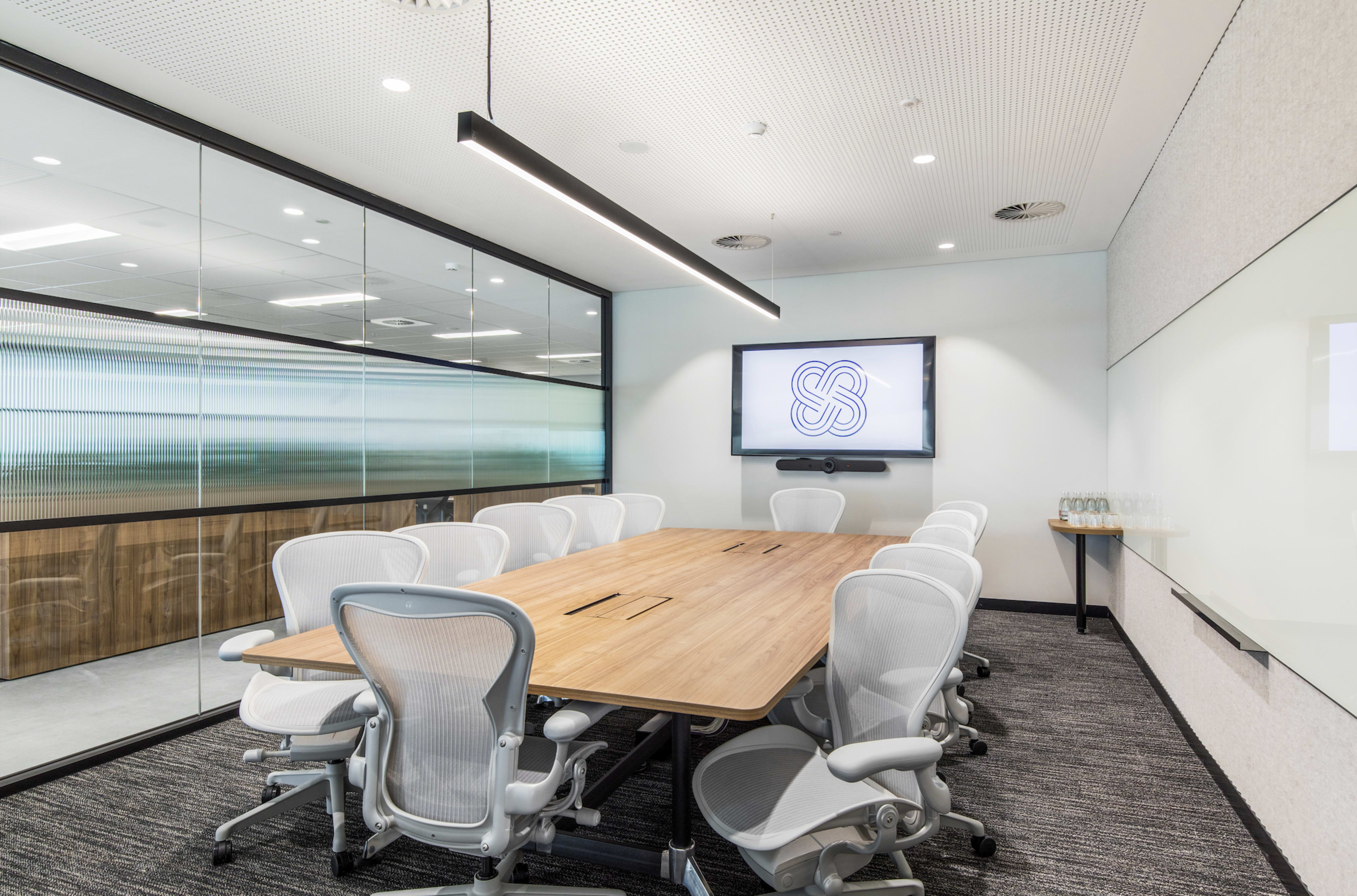 The image shows a modern conference room with a long wooden table, white ergonomic chairs, and a wall-mounted display.