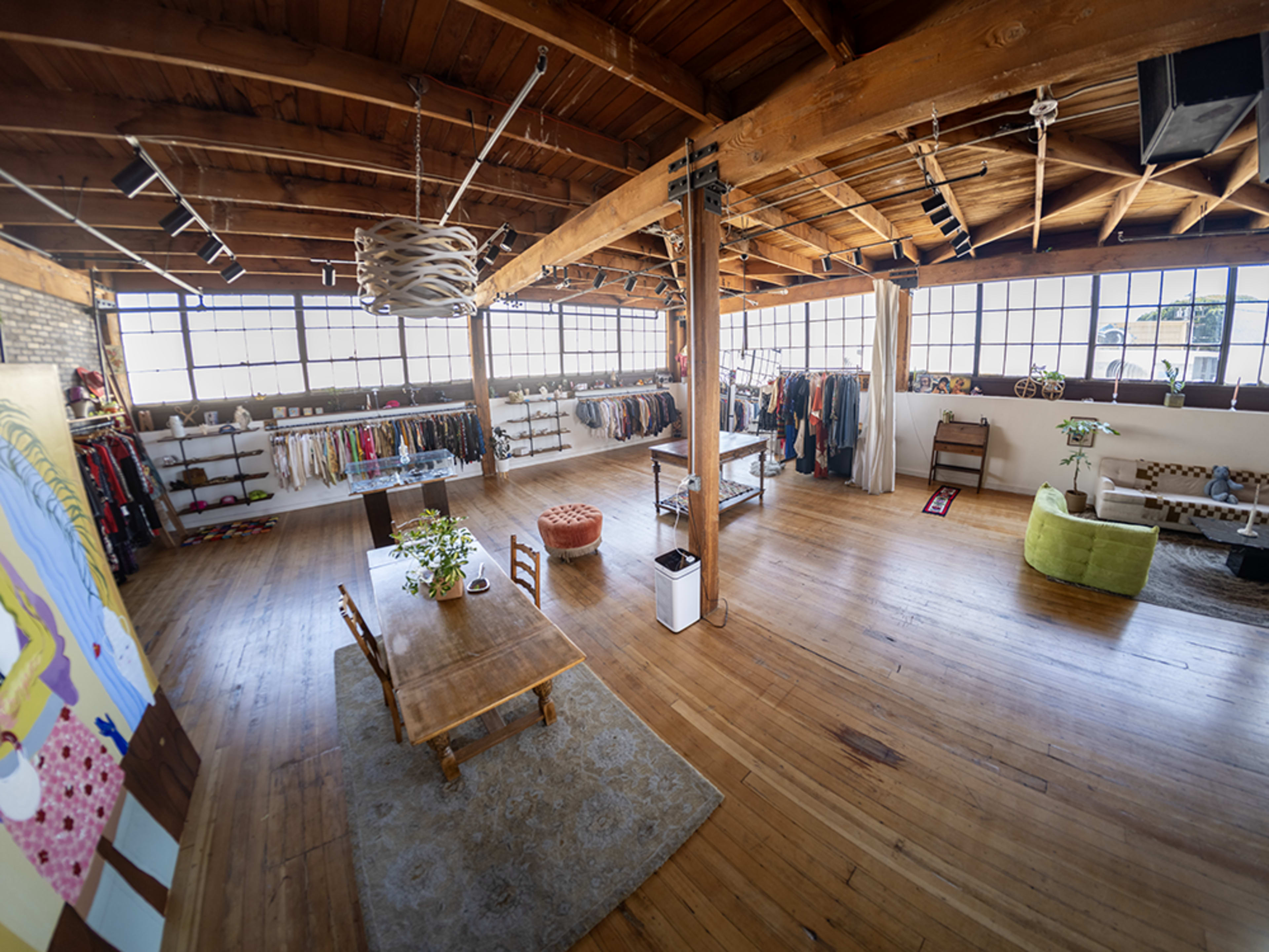 The image shows a spacious, well-lit room with wooden beams, featuring clothing racks along the walls, a dining table in the center, and a green sofa in the corner.