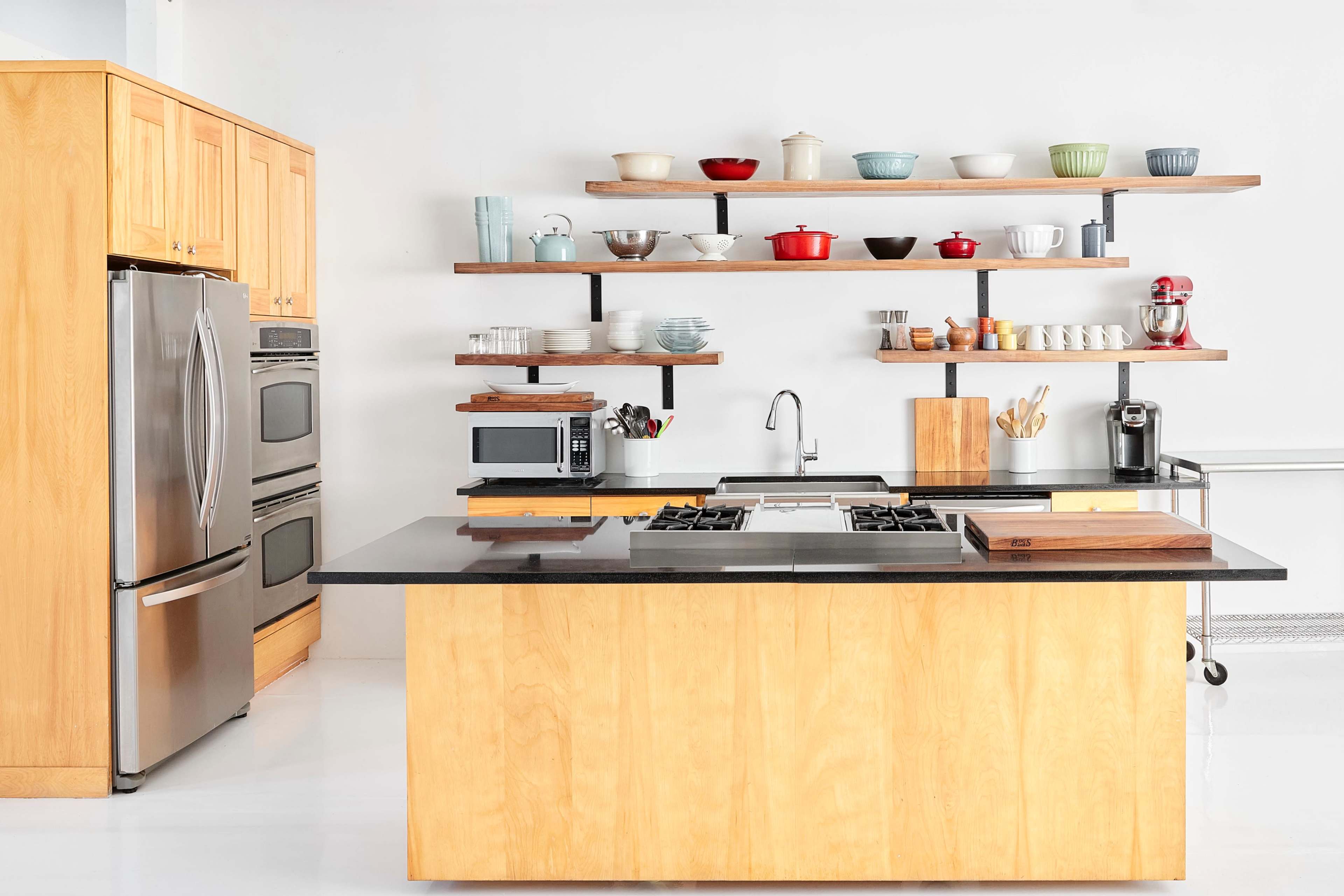 A modern kitchen features a central island with a black countertop, stainless steel appliances, and open shelving displaying various dishes and kitchenware.