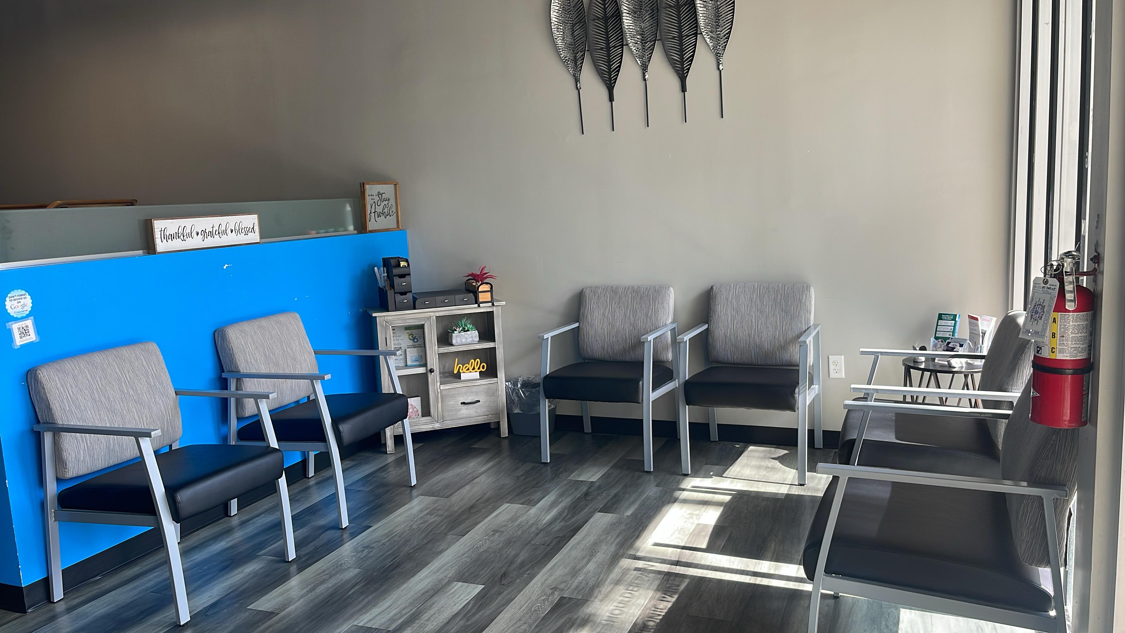 The image shows a waiting area with several gray chairs arranged around a small table, against a backdrop of light blue and gray walls.