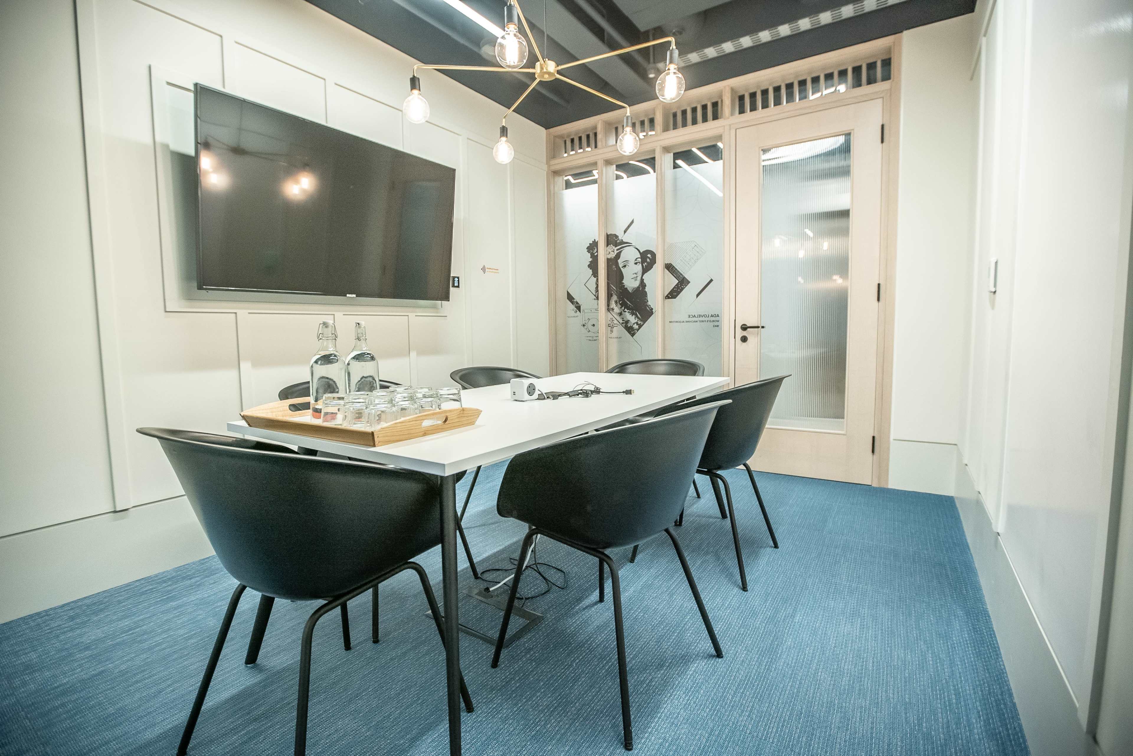 A modern conference room features a white table surrounded by black chairs, a large screen on the wall, and a door leading to another space.