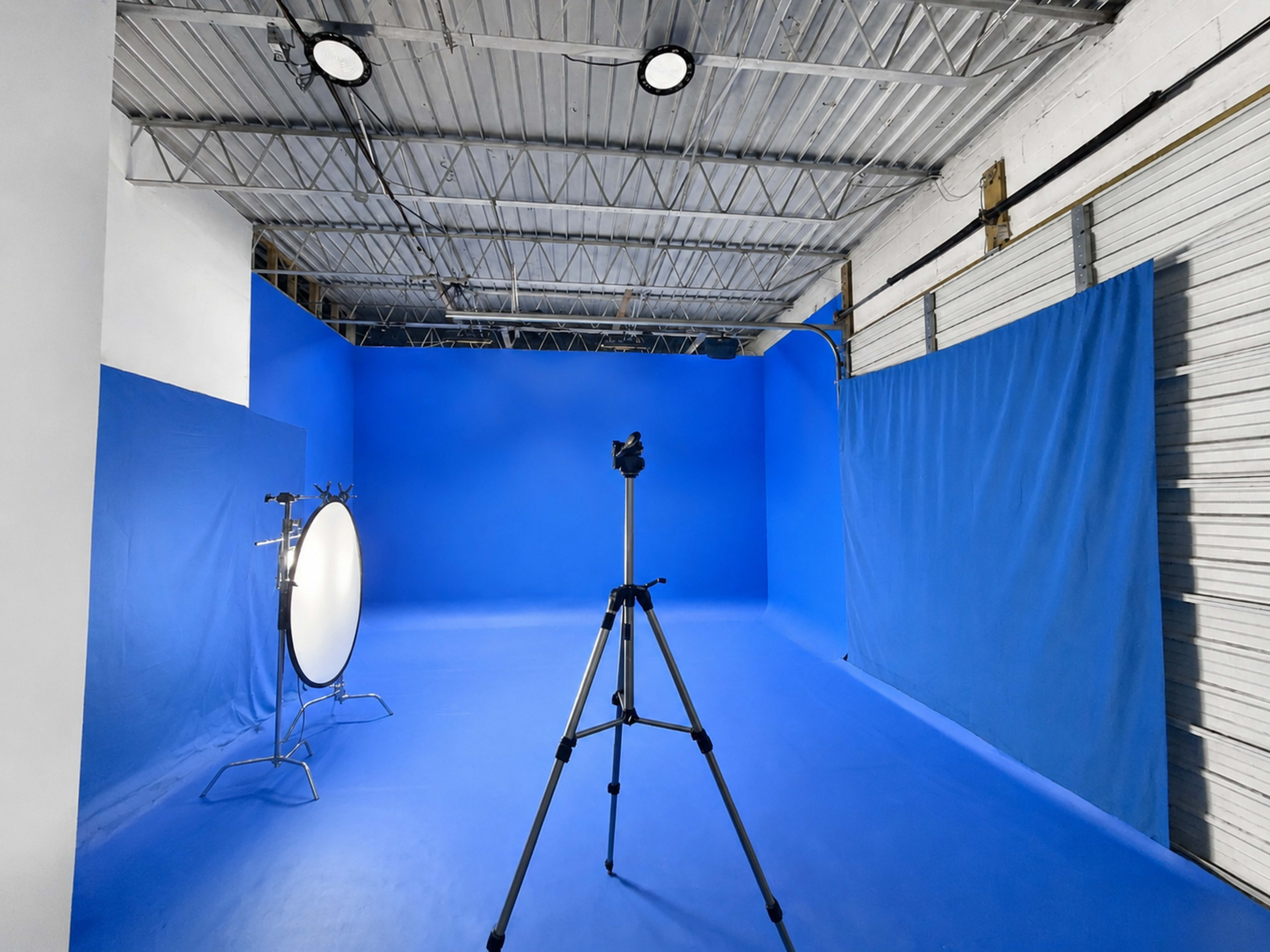 The image shows a photography studio with a blue backdrop, a tripod, and a reflector positioned on the floor.