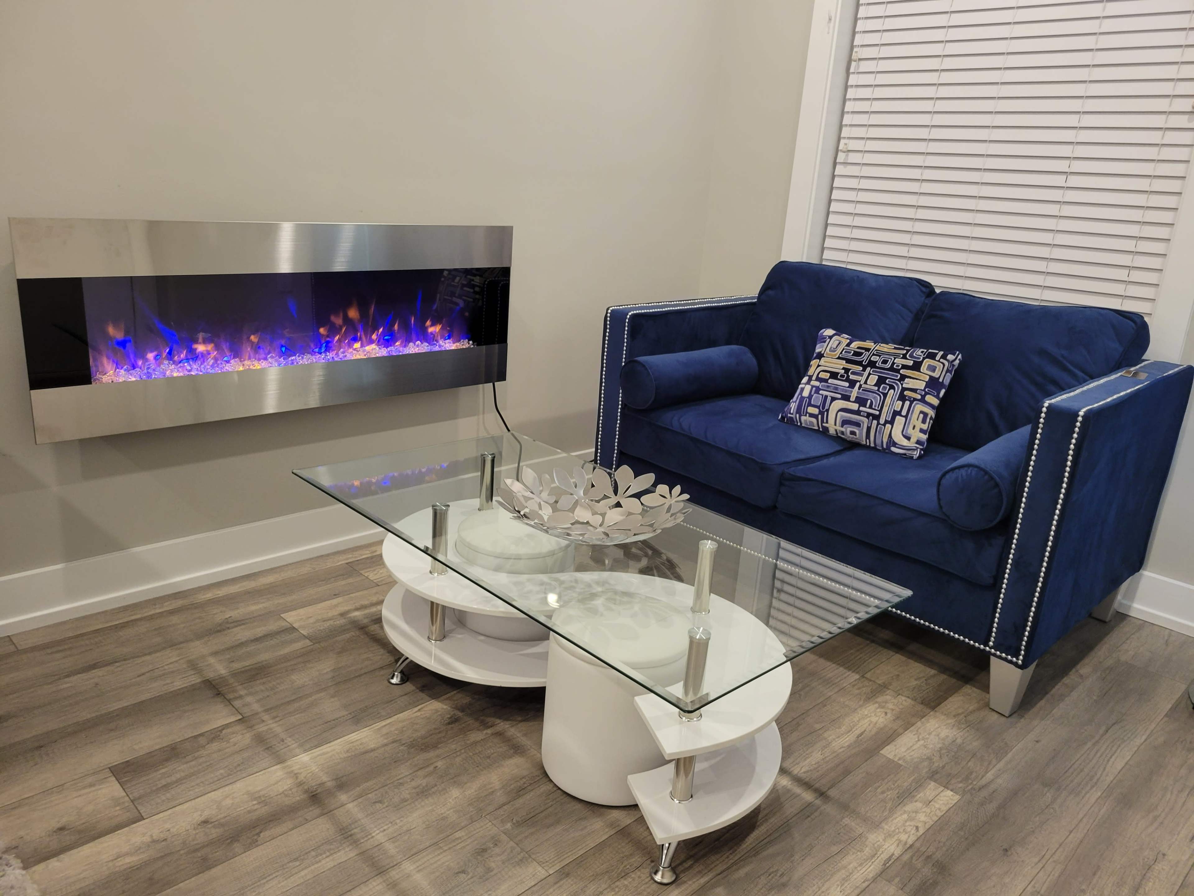 A modern living space featuring a blue sofa, a glass coffee table, and an electric fireplace mounted on the wall.