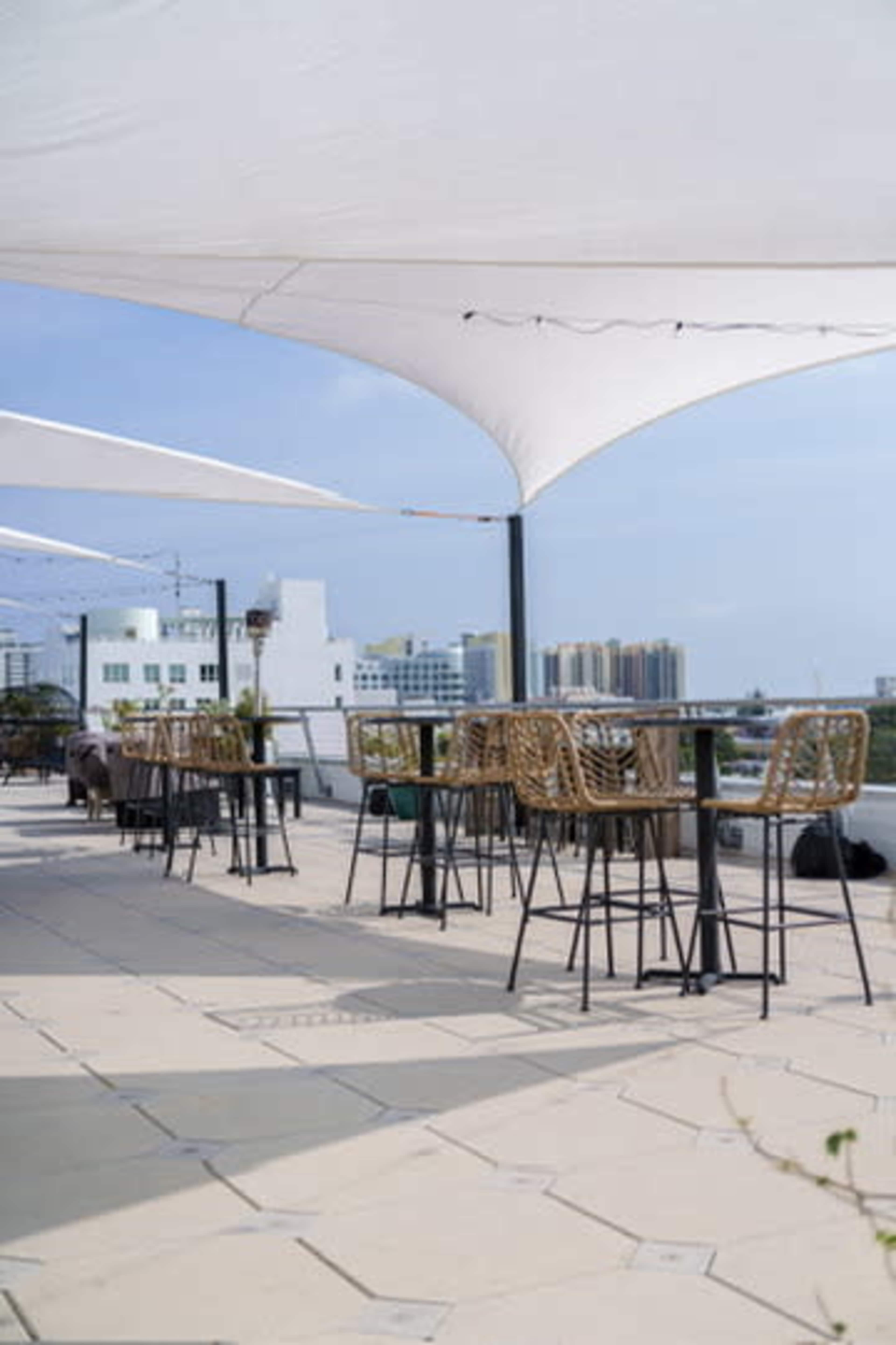 The image shows an outdoor rooftop terrace with tables and chairs under white canopies, overlooking a cityscape.